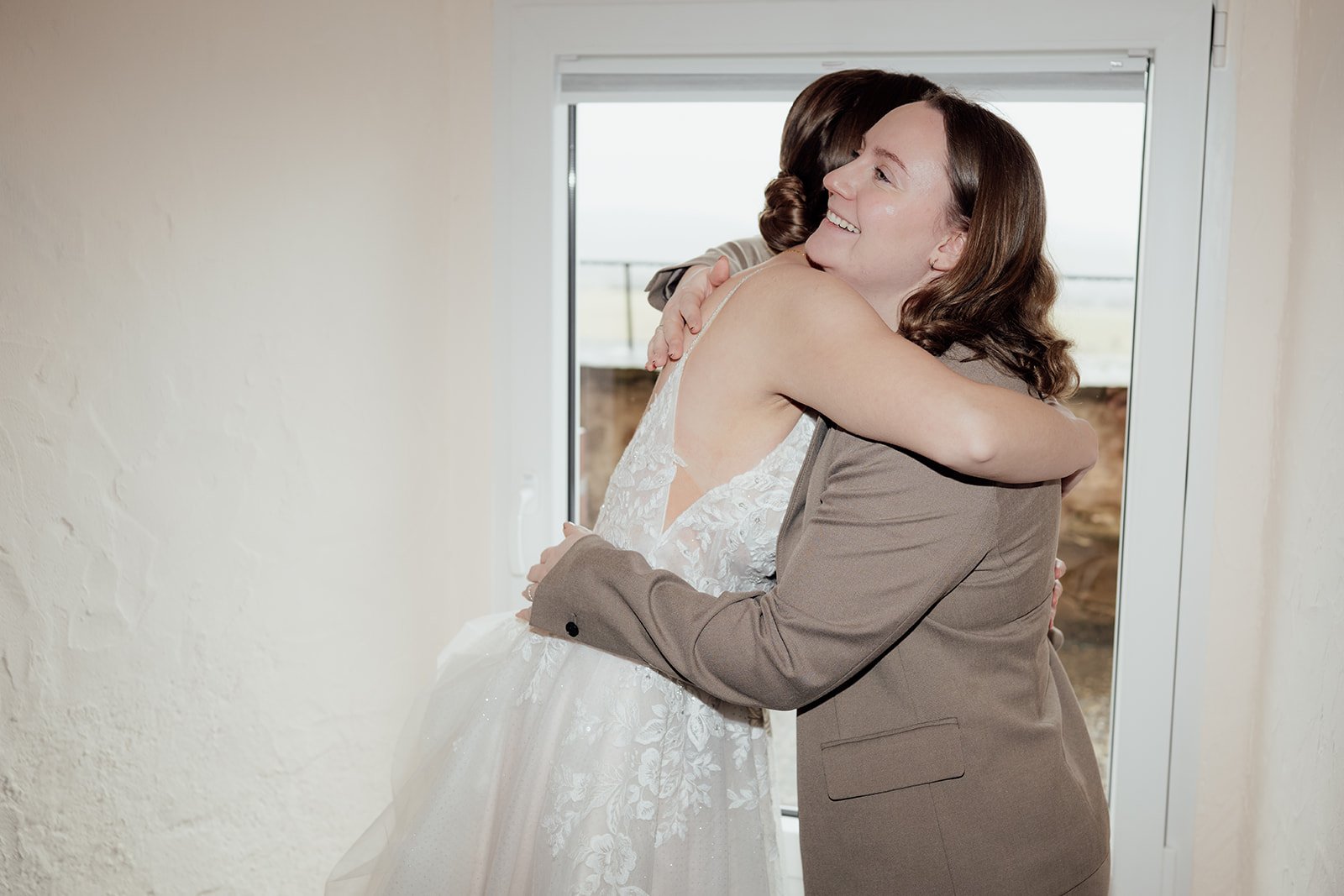 Eine Frau in einem Brautkleid umarmt eine andere Frau in einem brauen Anzug. Sie stehen dabei vor einem Fenster