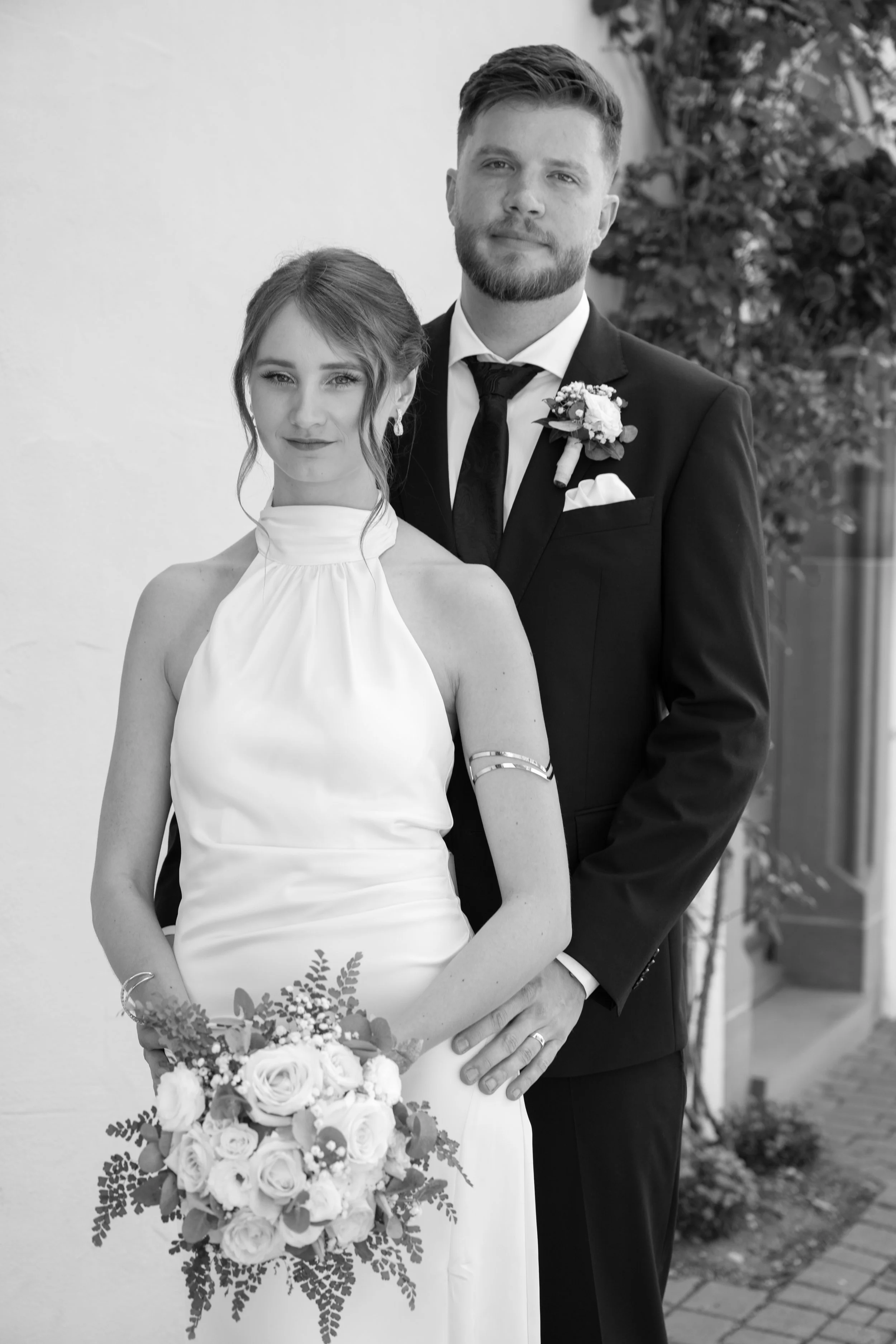 Black and white photo of a bride and groom, with the bride holding a bouquet of flowers, both dressed in formal wedding attire, standing outside against a plain wall.