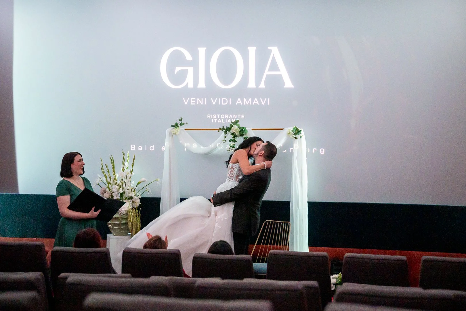 Weitwinkelaufnahme von einer Hochzeit in einem Kinosaal. Der Bräutigam hebt die Braut hoch und küsst sie