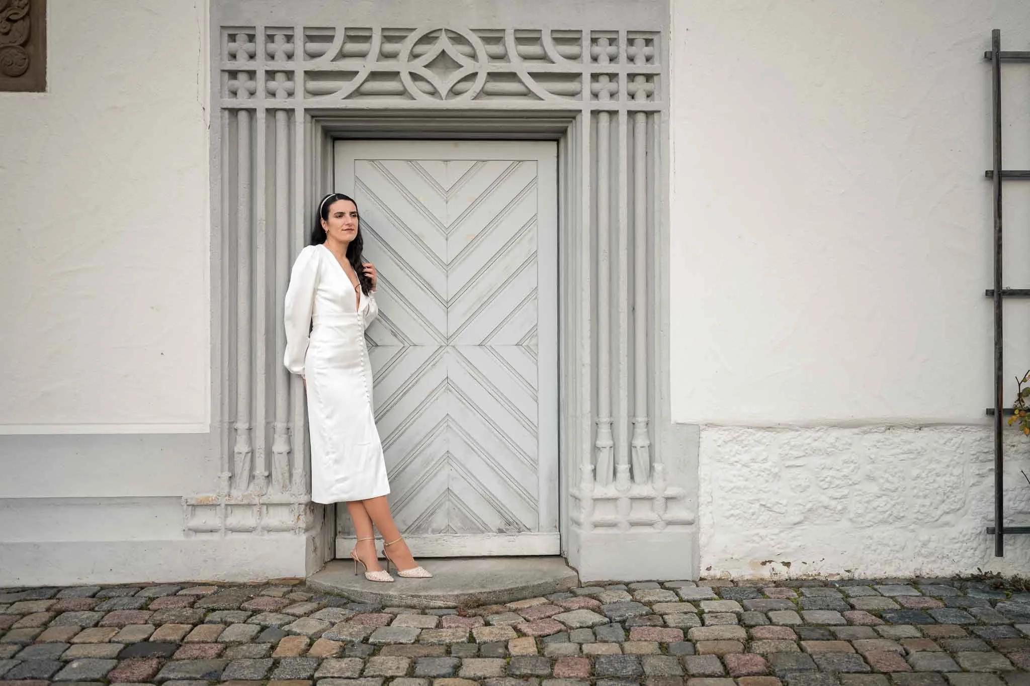 Eine Frau in einem kurzen Hochzeitskleid lehnt sich an den Türrahmen von einem historischen Gebäude an während sie in die Ferne blickt und sich dabei mit einer Hand über das Haar streicht