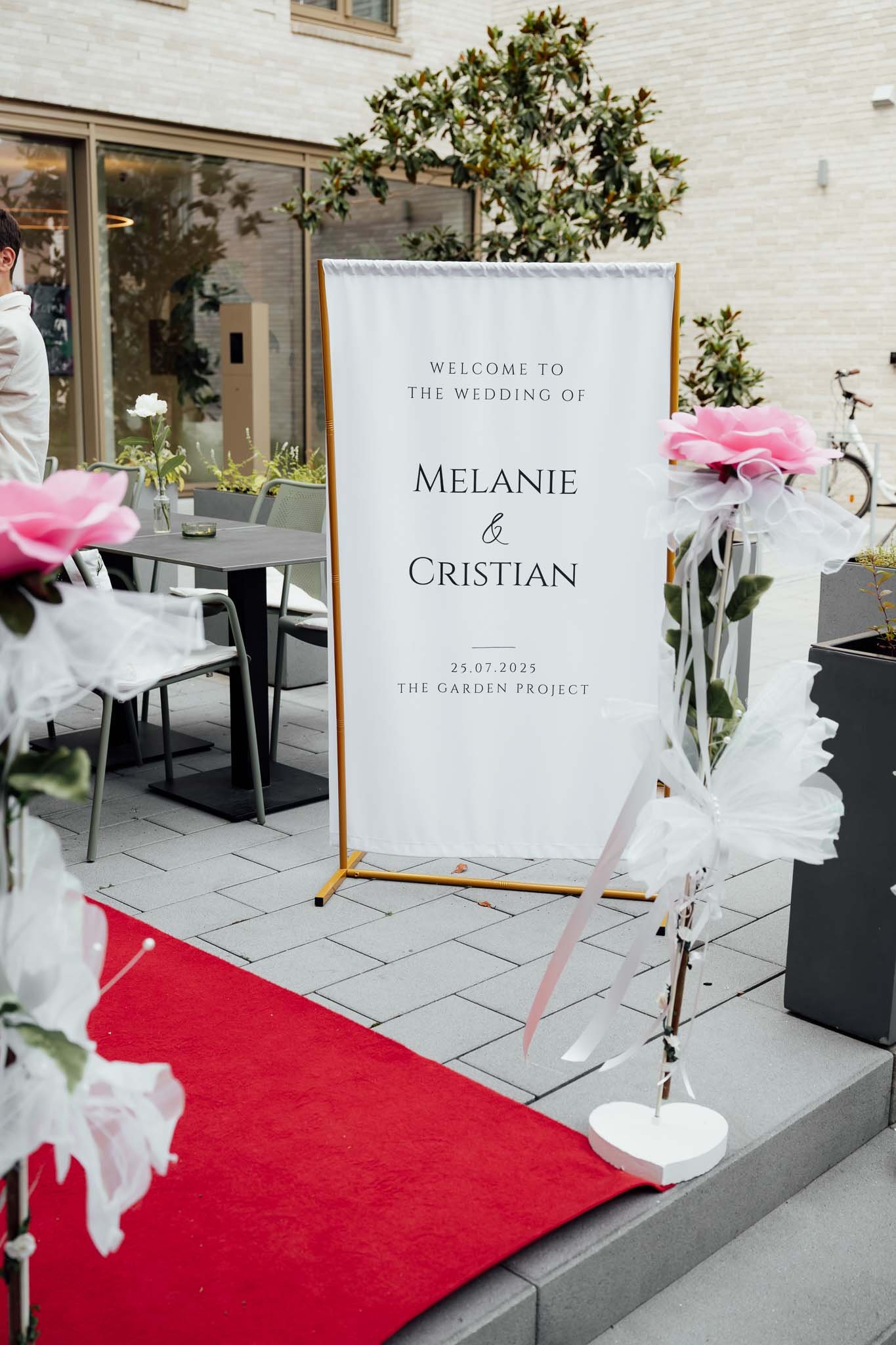 Willkommensschild zur Hochzeit von Melanie und Cristian, Datum 25.07.2025, im Gartenbereich, mit roten Teppich und Dekoration aus weißen und pinken Blumen.