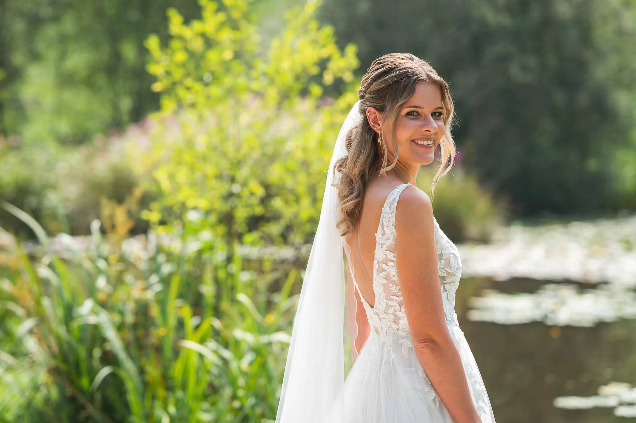 Lächelnde Braut in einem weißen Brautkleid mit Spitzenärmeln im Grünen am Wasser.