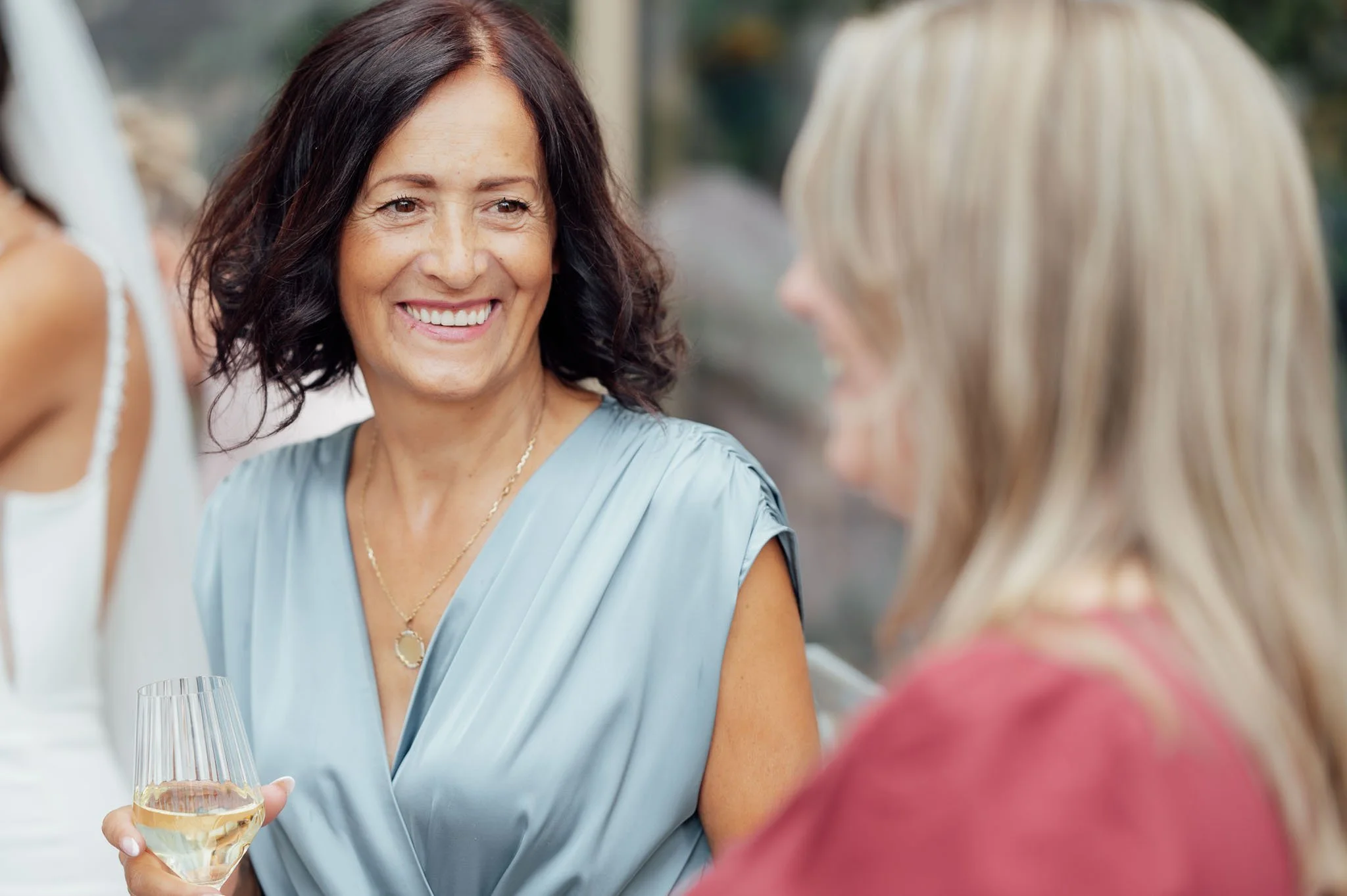Zwei Frauen, eine mit dunklen Haaren in einem blauen Kleid und eine mit blonden Haaren in einem roten Kleid, beim Gespräch bei einem festlichen Anlass, eine hält ein Glas Weißwein.