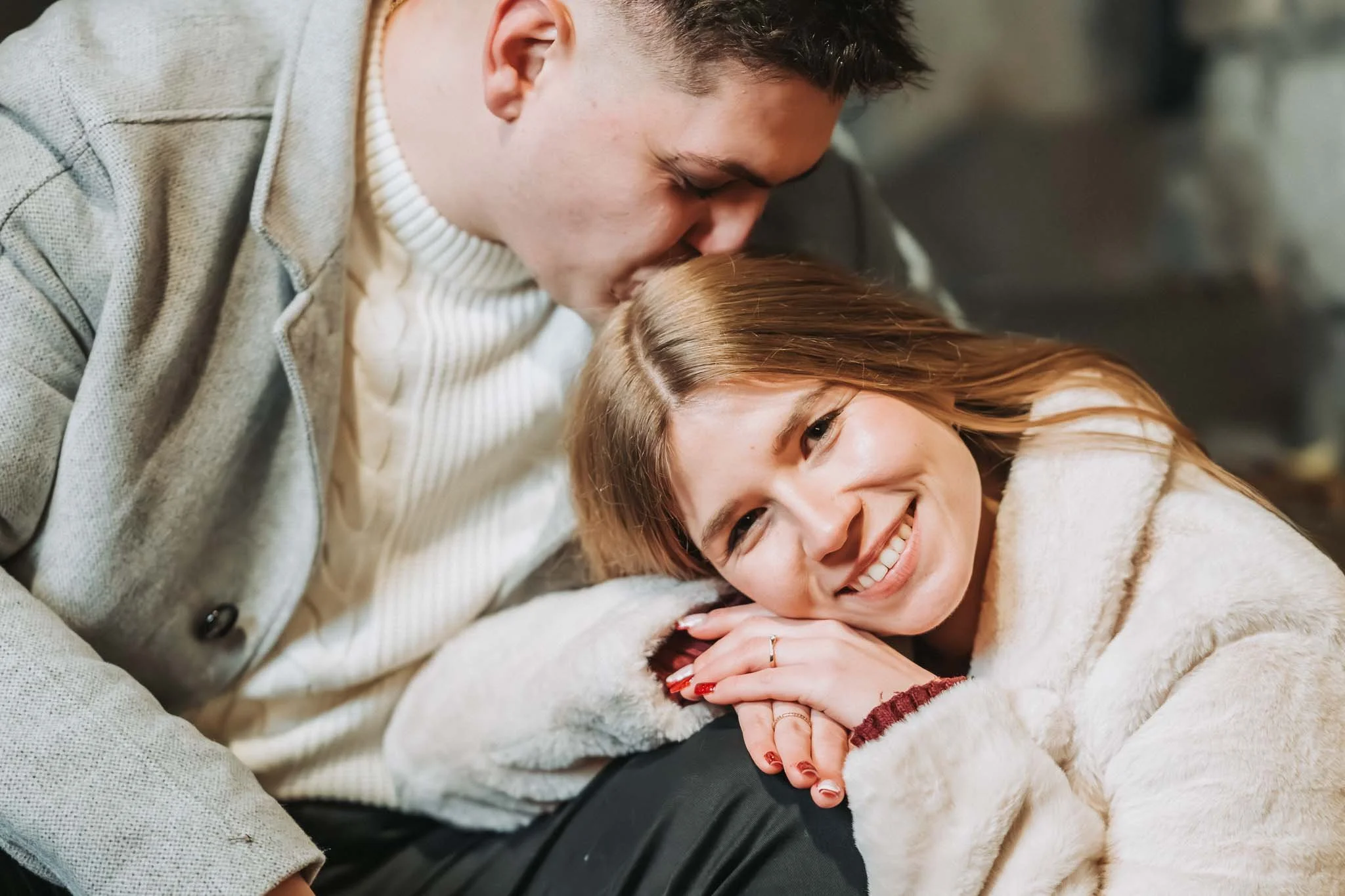 Eine Frau ist mit dem Kopf auf das Knie eines Mannes angelehnt, der ihr einen Kuss auf die Stirn ibt. Beide lachen glücklich. Sie tragen Mäntel.