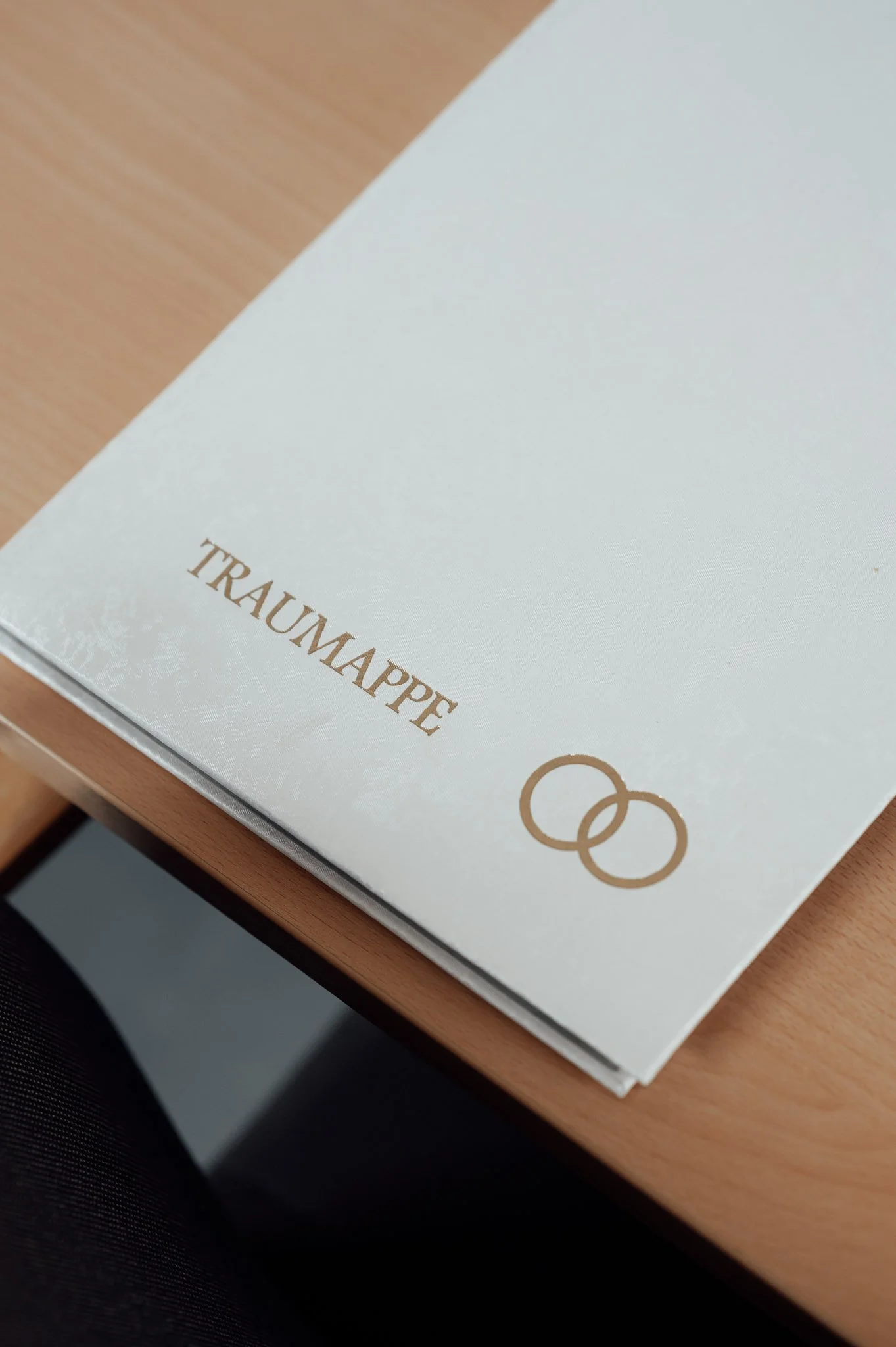Ein weißes Buch oder Dokument mit den Worten "Traumapp" und zwei überlappenden Ringen in Gold auf dem Cover, liegt auf einem Holztisch.