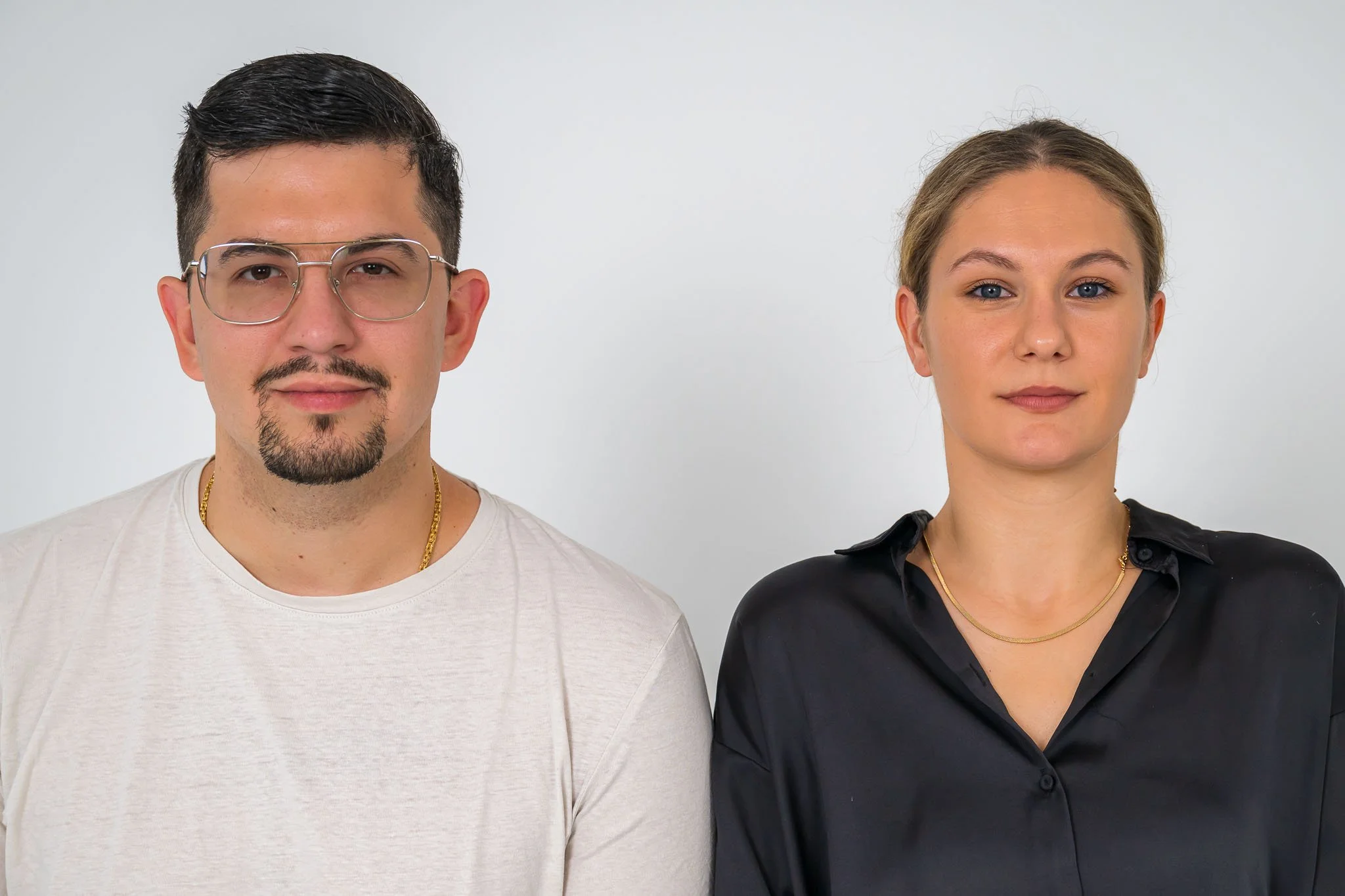 Ein Mann im weißen Shirt sitzt vor der Kamera, seine Schulter berührt eine Frau, die ein schwarzes Hemd trägt, die aber identisch wie er in die Kamera blickt.