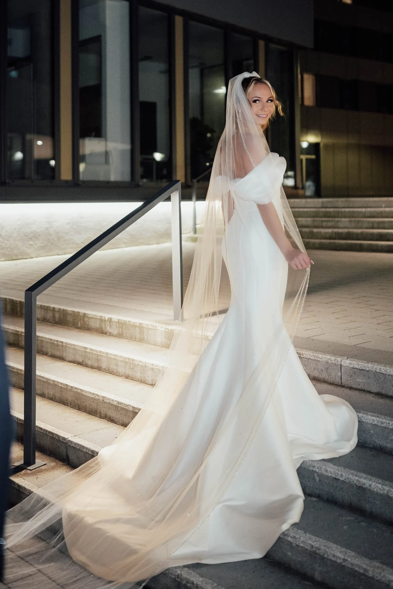 Eine Braut in einem weißen Hochzeitskleid mit Schleier vor einem modernen Gebäude bei Nacht.