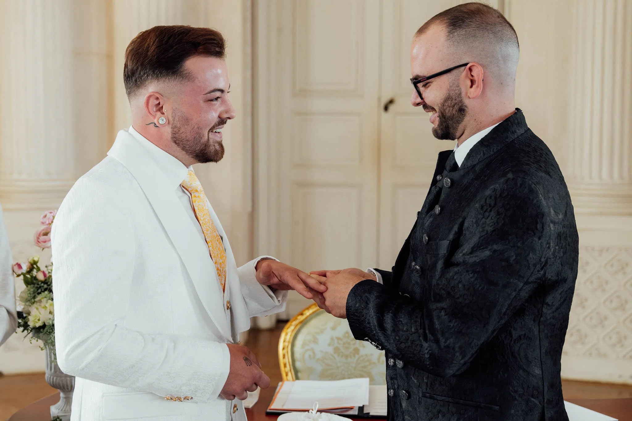 Zwei Männer bei einer gleichgeschlechtlichen Hochzeit, die sich die Hände halten und lächeln, in einem eleganten Raum mit Blumen.