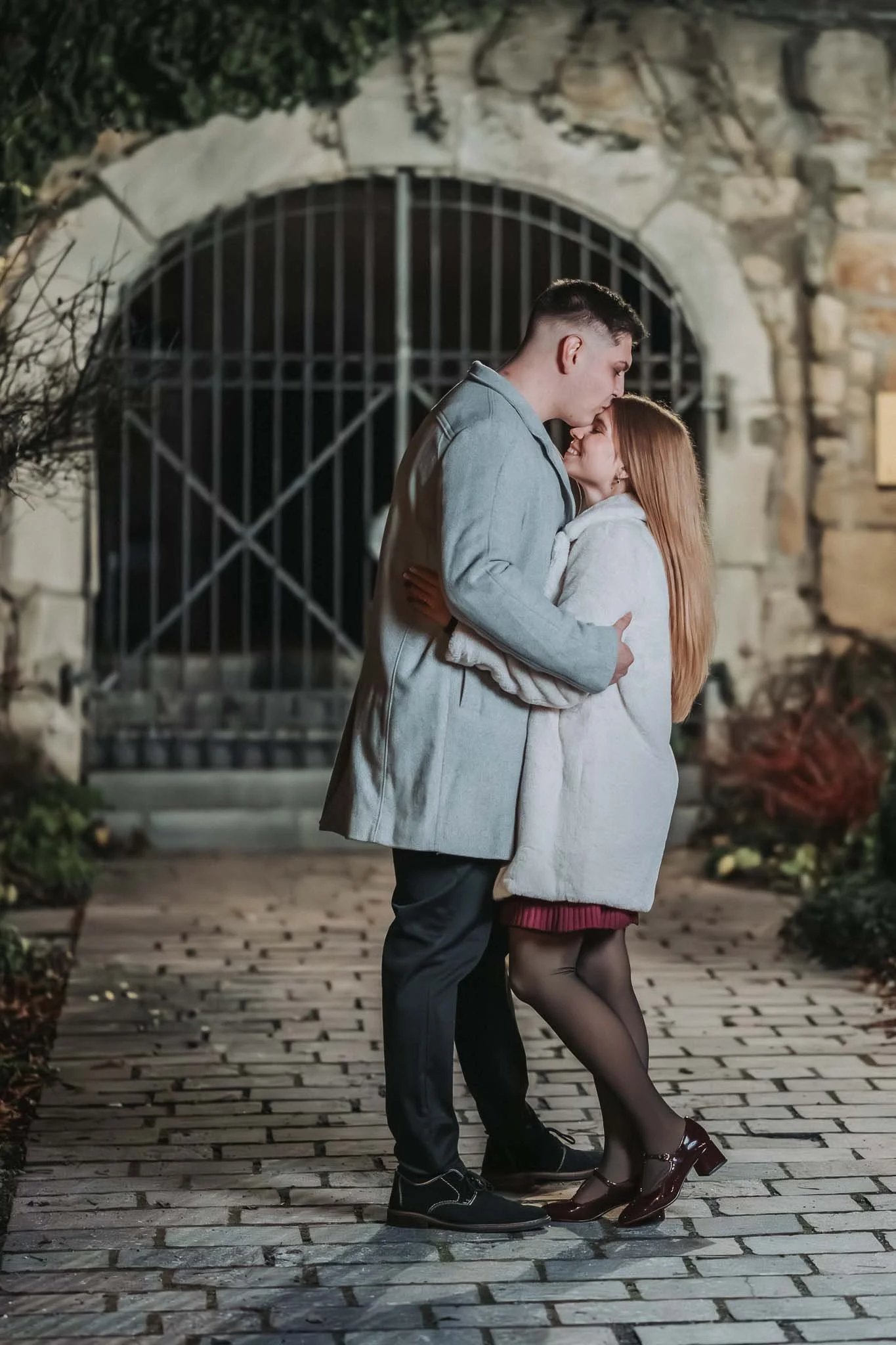 Eine Frau und ein Mann stehen vor dem Tor einer Steinmauer. Er gibt ihr einen Kuss auf die Stirn, sie hat die Augen geschlossen. Beide sind winterlich angezogen.