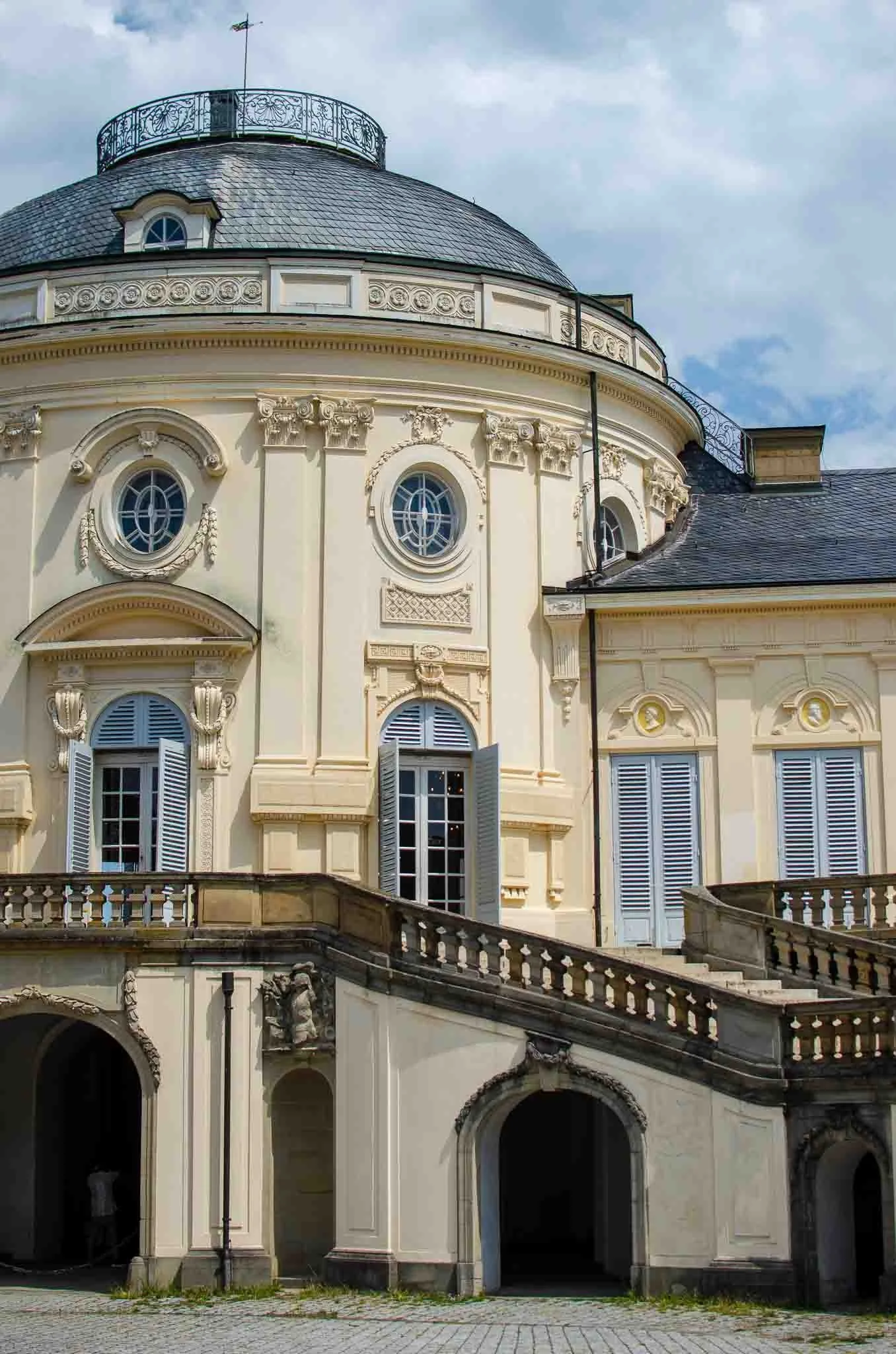 Ein barockartiges Gebäude mit einer großen Kuppel, geschmückten Fassaden und mehreren Fenstern mit Holzläden, teilweise offen, vor wolkigem Himmel.