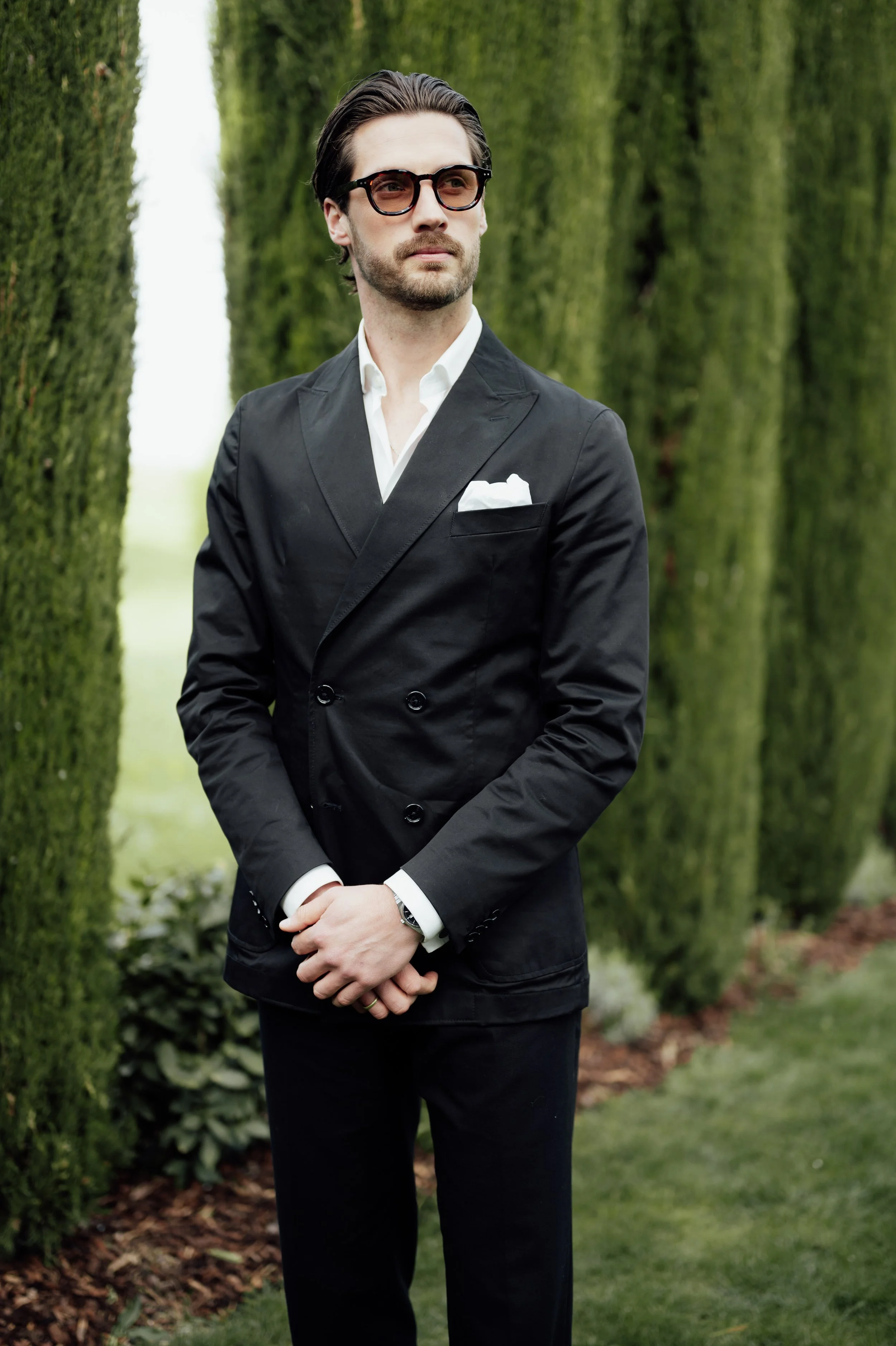 A man wearing glasses and a black suit with a white dress shirt and pocket square standing outdoors in front of tall, green hedge trees. His hands are folded in front of him.