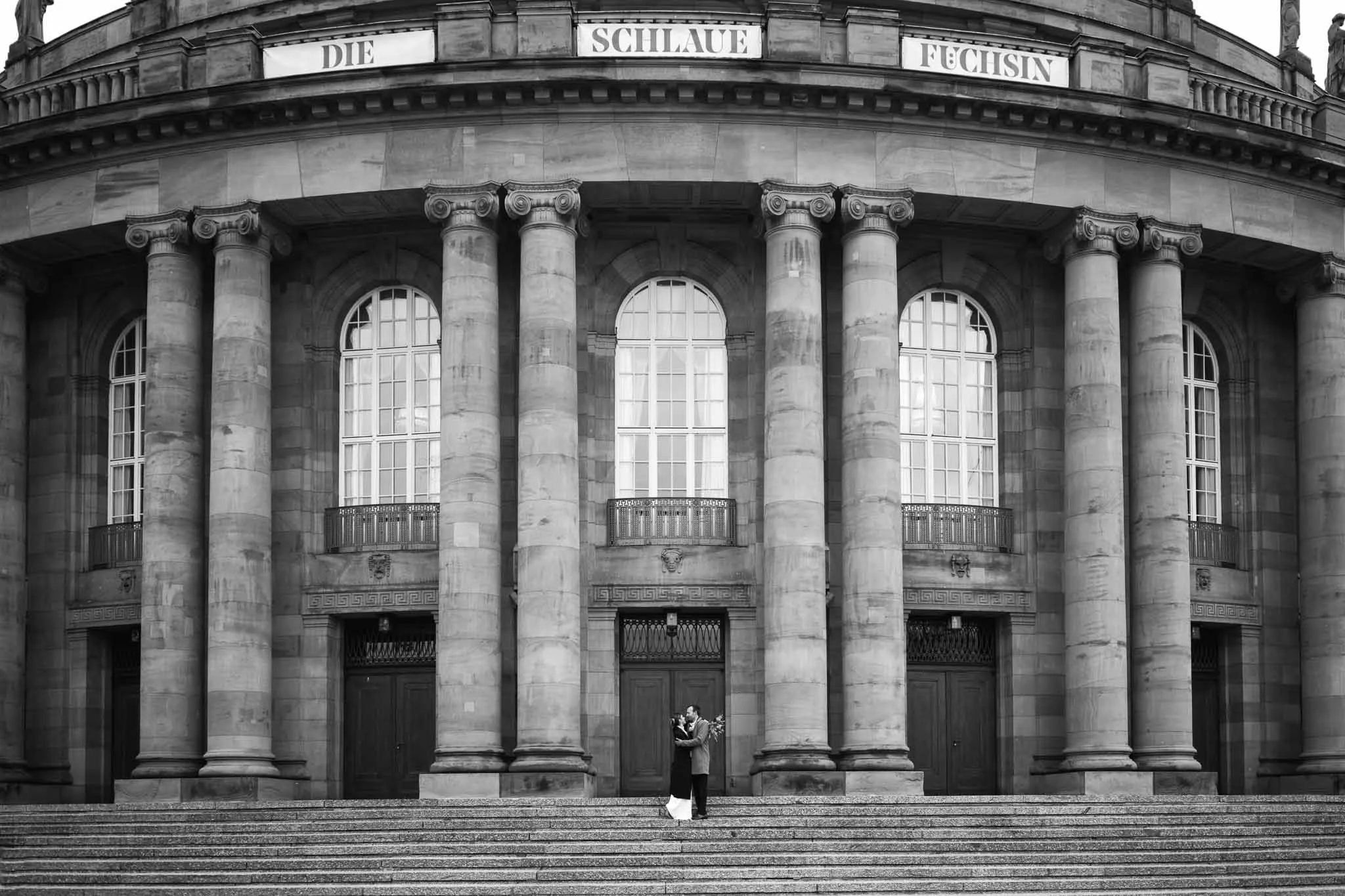 Ein Brautpaar steht auf den Treppen vor einem großen, klassischen Gebäude mit Säulen und großen Fenstern, die auf Deutsch als 'Die Schlauen Füchsin' bezeichnet werden.