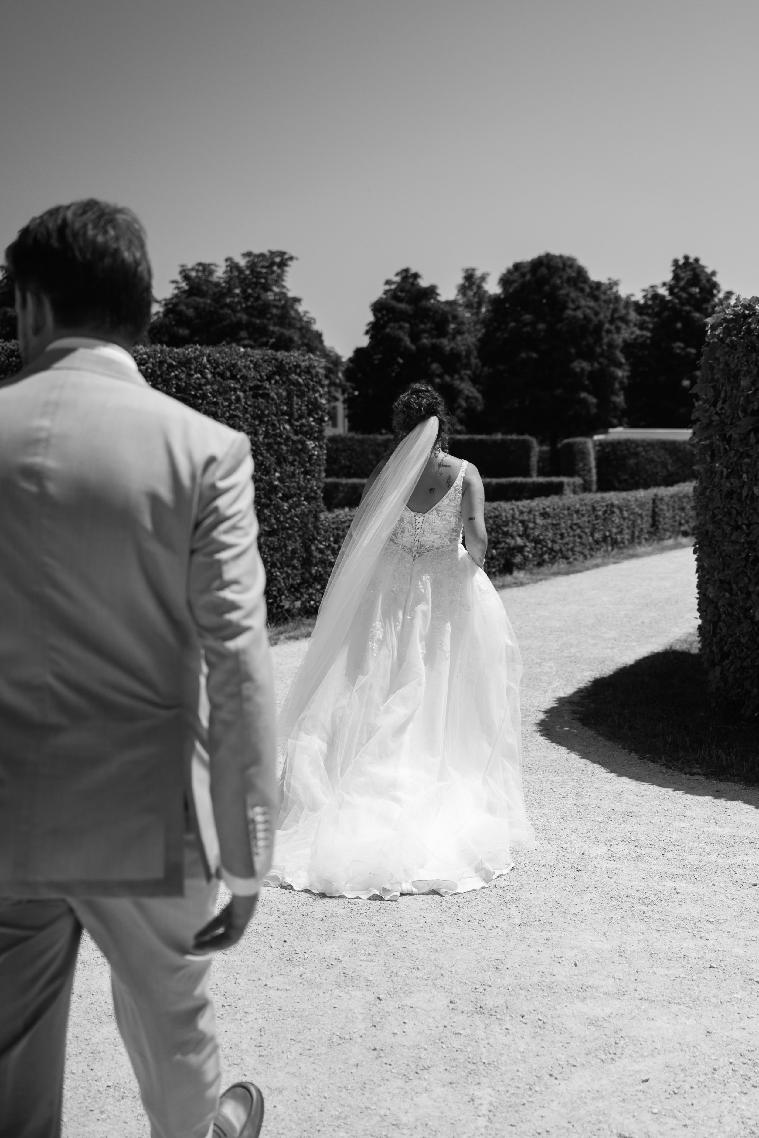 A wedding scene with a bride in a white gown and veil walking away, seen from behind, and a man in a light-colored suit walking towards her, on a pathway with manicured hedges and trees in the background.