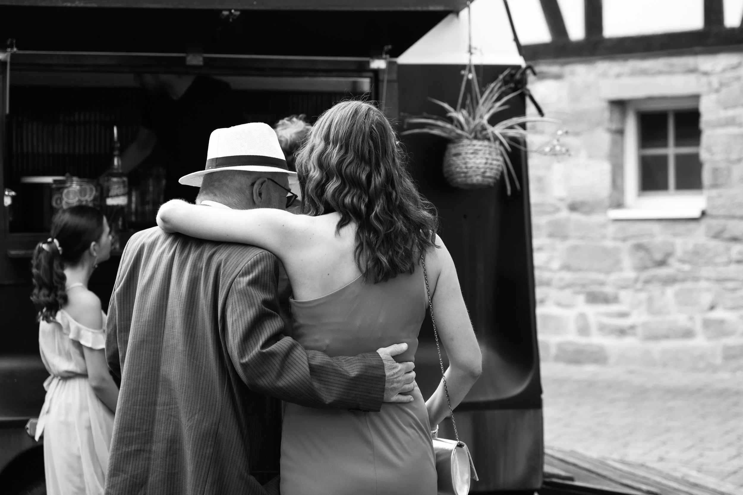 Black and white photo of an elderly man and a woman embracing each other at a social gathering. The man is wearing a hat and glasses, while the woman has curly hair. There are other people in the background and a brick building.