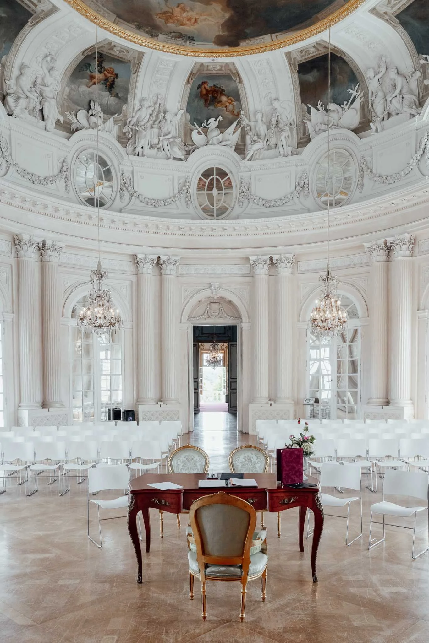 Innenraum eines prunkvollen, weißen Barocksaals mit hohen Fenstern, Kristallleuchtern, verzierten Säulen und einer bemalten Kuppeldecke, im Vordergrund ein roter Schreibtisch mit Stühlen und Blumen.
