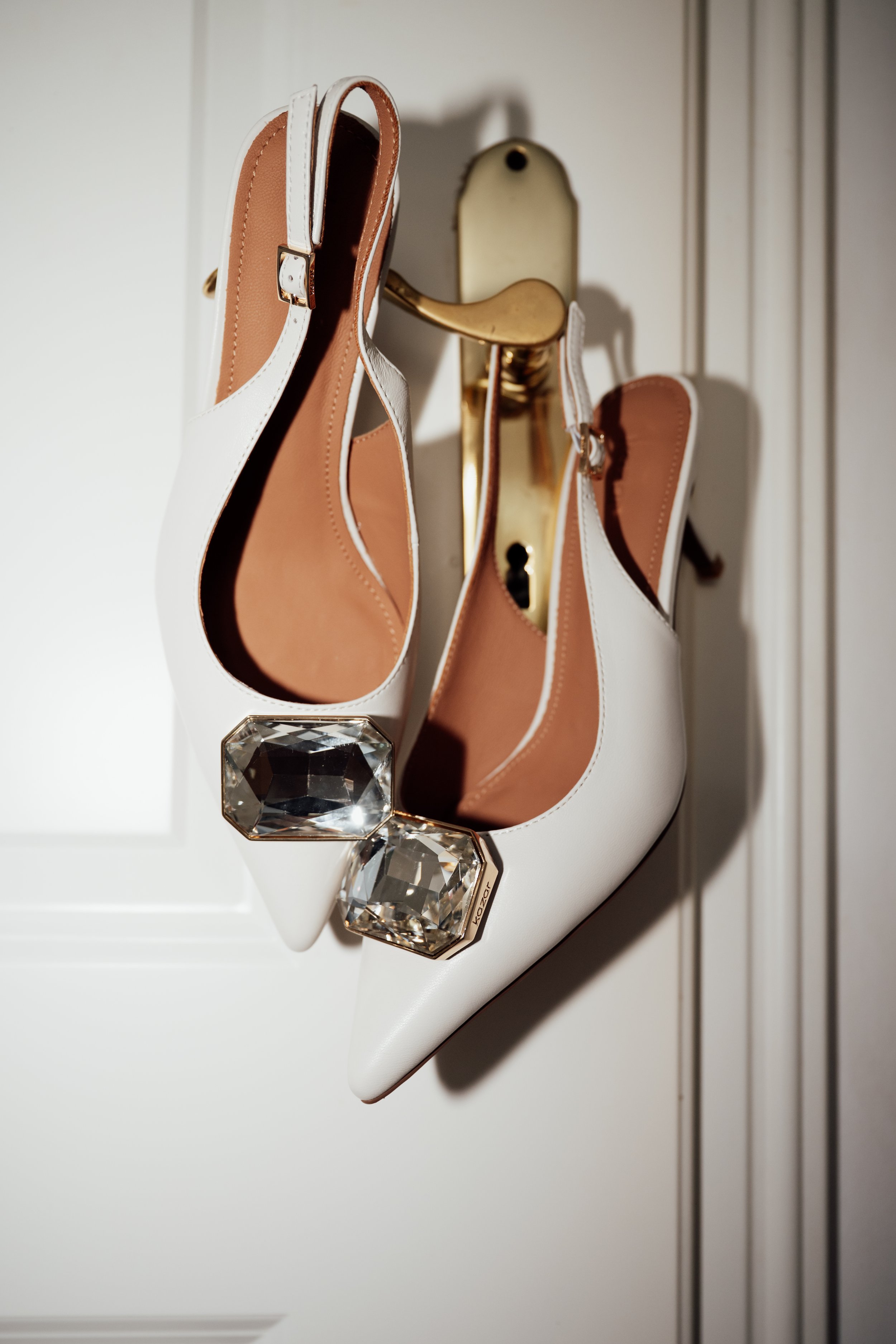 A pair of white high-heeled shoes with decorative large gemstone accents on the heels, hanging on a gold-colored hook on a white door.