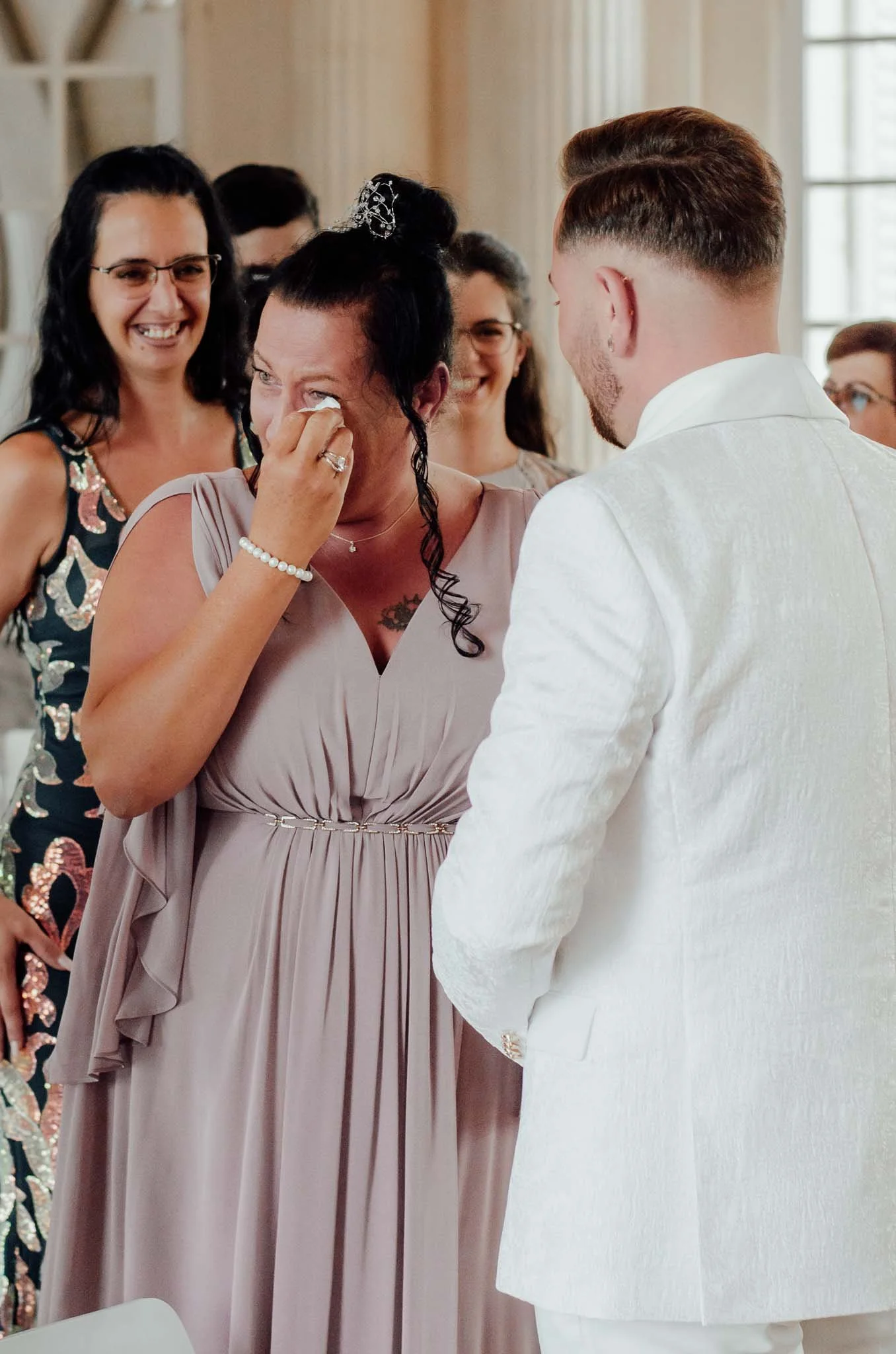 Eine Frau in einem Kleid weint, während sie an einer Hochzeit mit einem Bräutigam in einem weißen Anzug teilnimmt, umgeben von lachenden Damen in einem festlichen Raum.