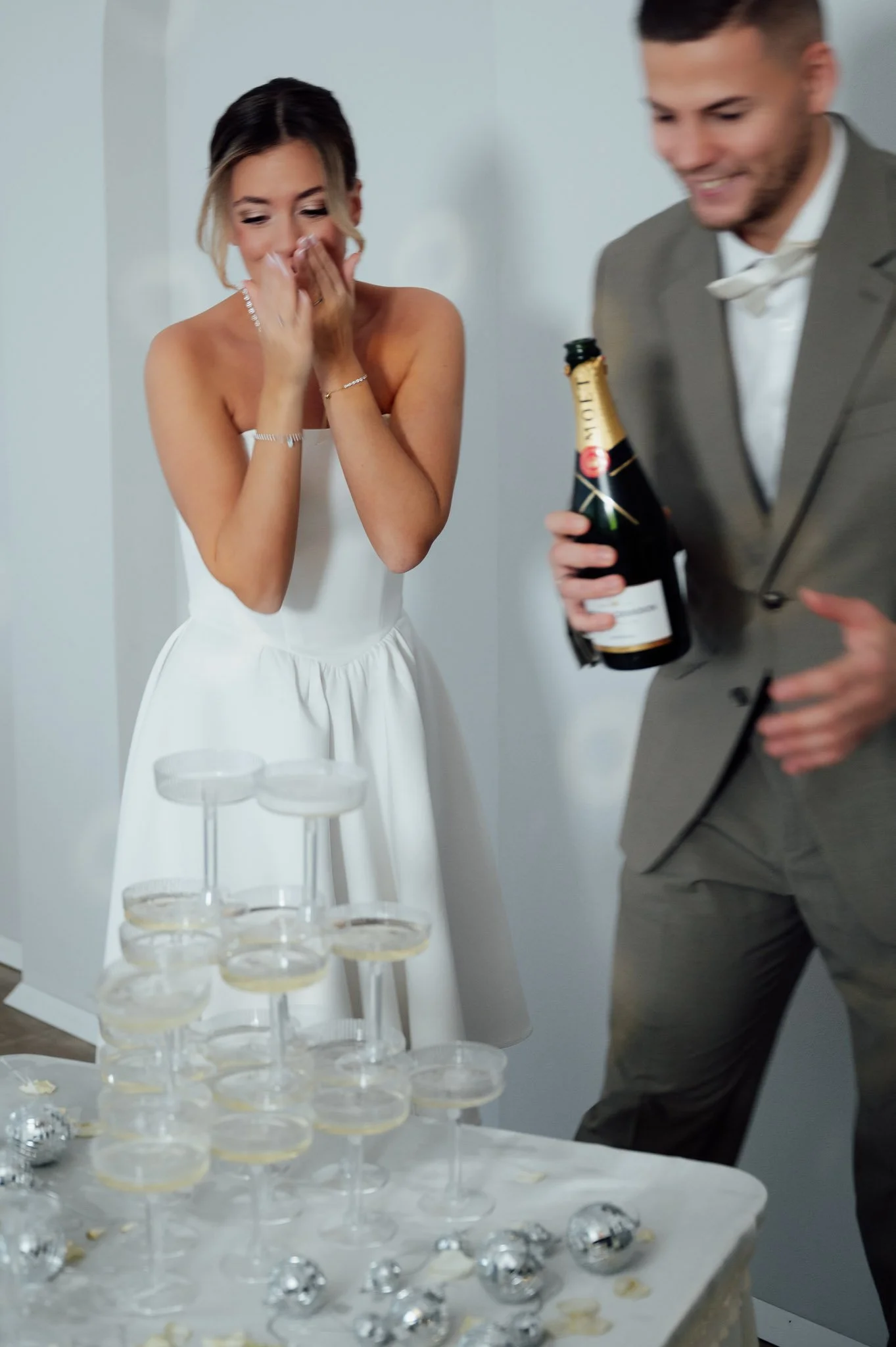Ein Mann in Anzug hält eine Flasche Sekt, während eine Frau in Brautkleid sie lachend und schmunzelnd betrachtet beim Feiern.