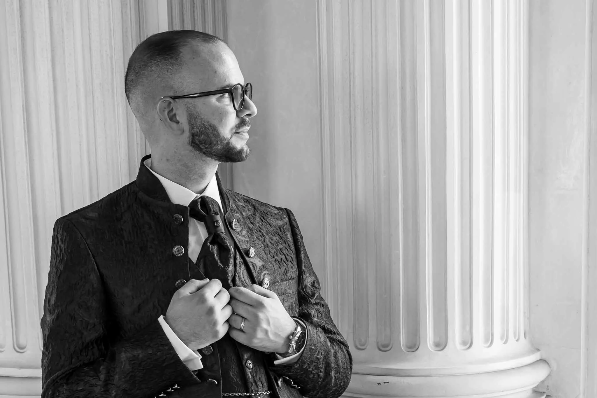 Ein Mann in traditioneller schwarzer Tracht mit Brille, posiert vor einer klassizistischen Säule, in Schwarz-Weiß aufgenommen.