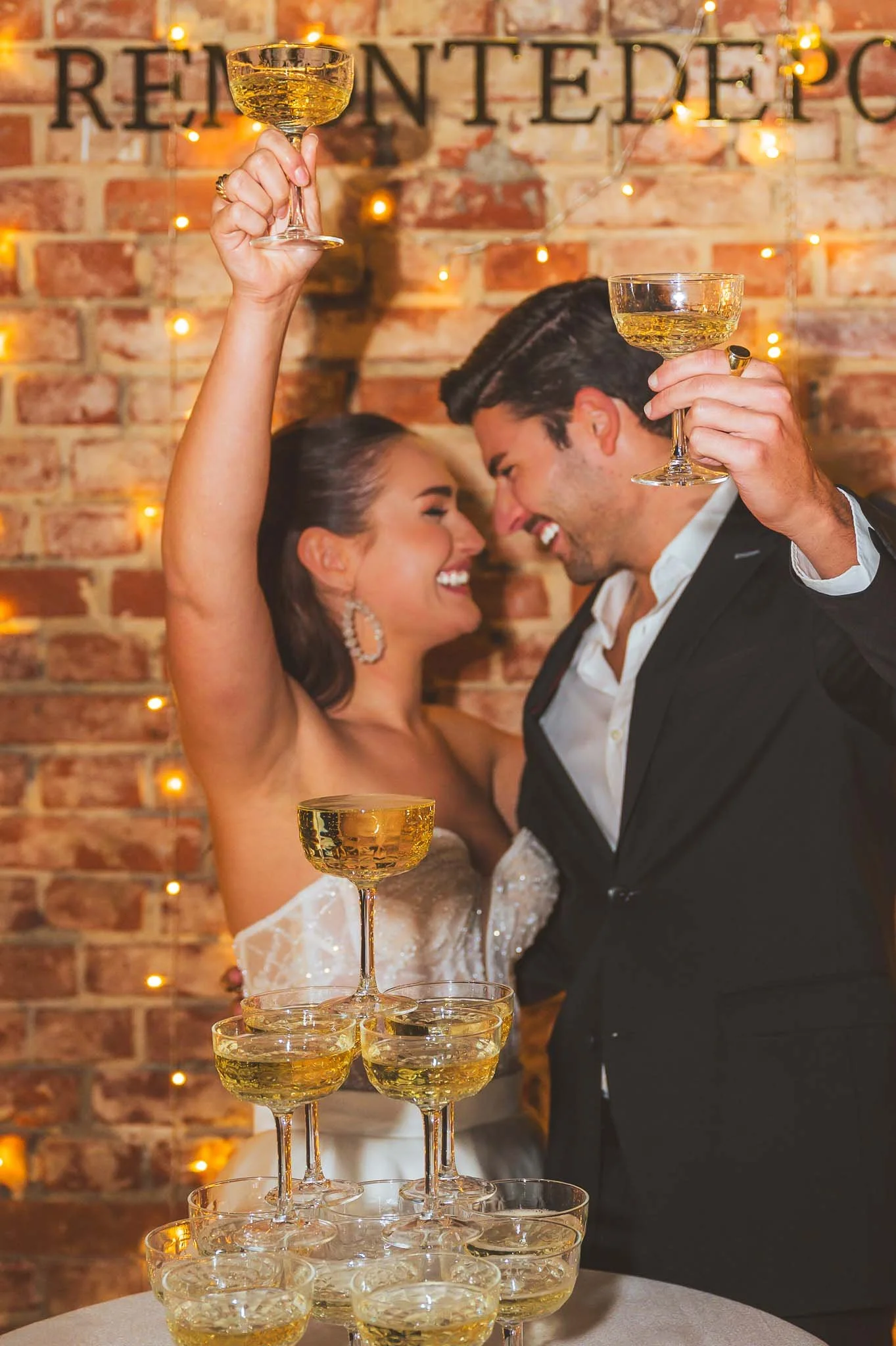 Ein feierndes Paar bei einer Hochzeit, das mit Champagner auf eine erfolgreiche Feier anstößt, vor einem Brickwall mit Lichterdeko.