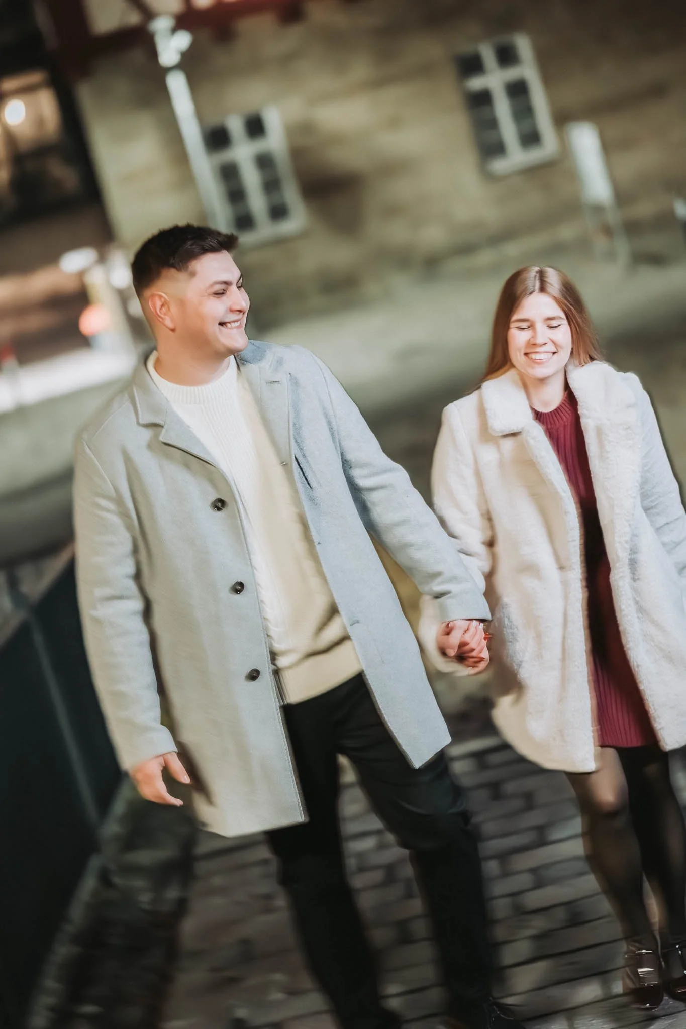 Verschwommenes Bild von einem Paar, welches sich an den Händen hält und lachend der Kamera entgegen läuft. Beide tragen Mäntel und die Frau trägt einen kleinen Blumenstrauß
