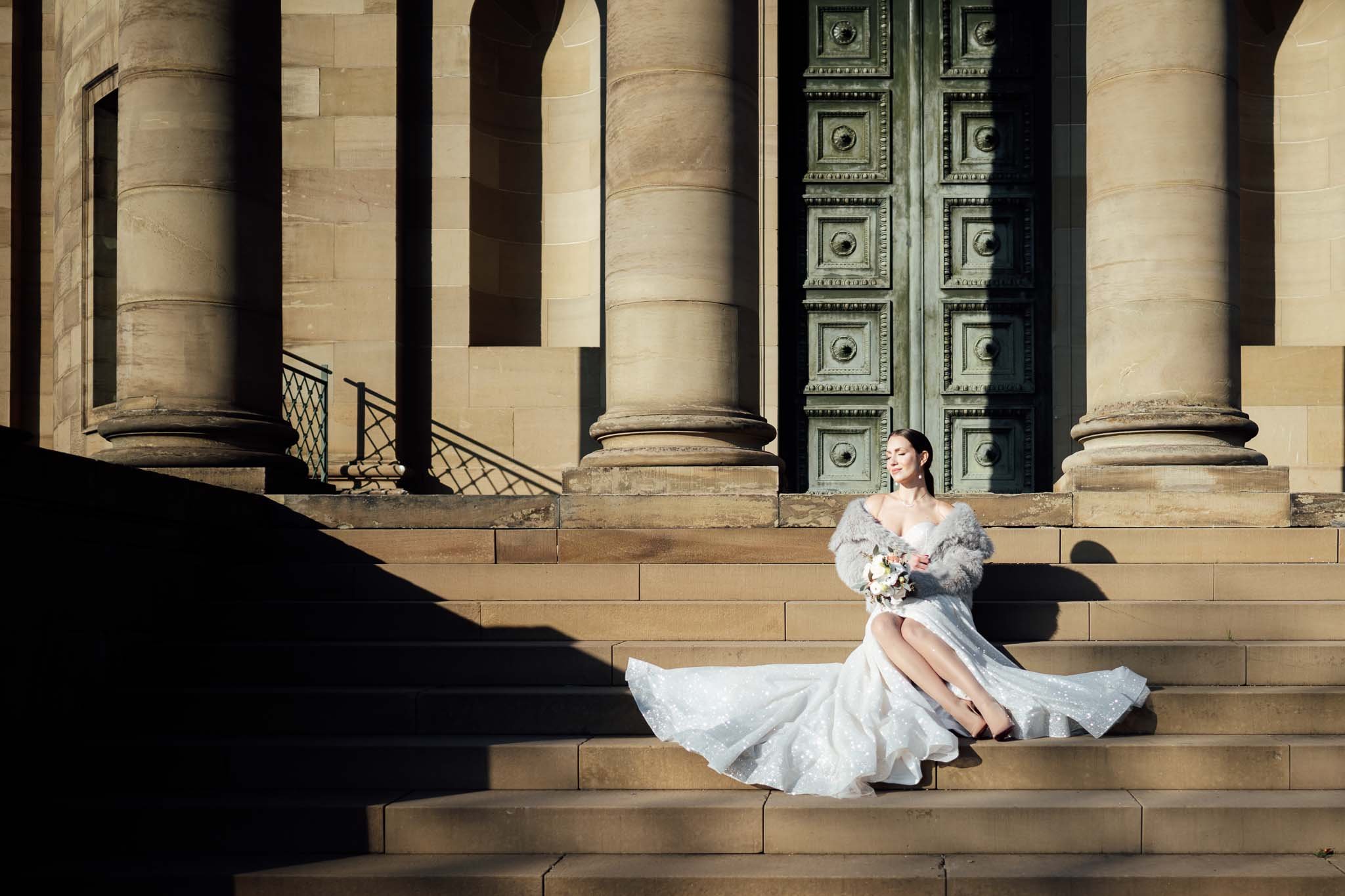 Brautpaarshooting an der Grabkapelle Stuttgart. Braut in weißem Brautkleid mit Schleier sitzt auf den Stufen einer historischen Kathedrale mit großen Steinsäulen und eine massive grüne Tür im Hintergrund, Sonnenlicht und Schatten.