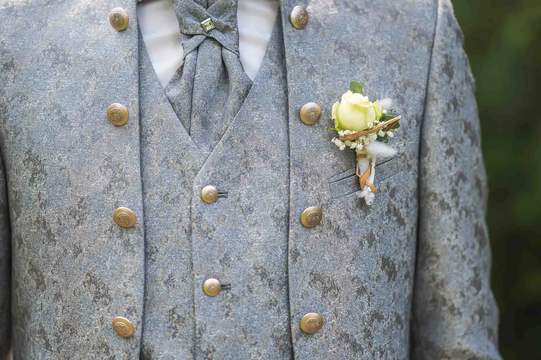 Nahaufnahme eines stilvollen männlichen Anzugs in Grau mit goldenen Knöpfen, mit einem weißen Blumen- und Federbroschen an der Brusttasche.