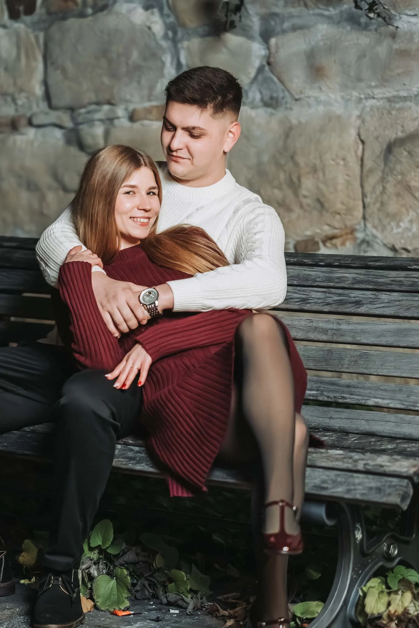 Ein Mann sitzt auf einer Parkbank und eine Frau liegt in seinen Armen. Sie lächelt in die Kamera, während er sie ruhig ansieht.