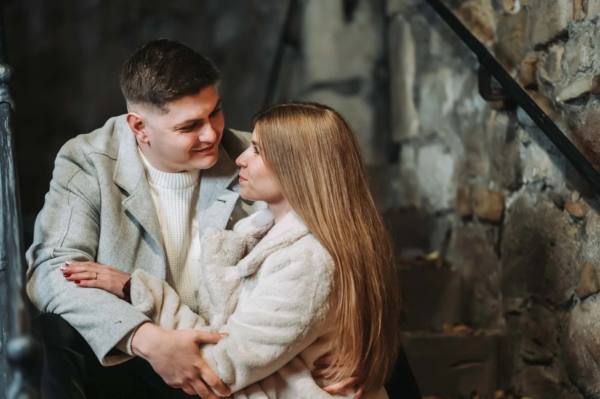 Ein Paar sitzt auf einer Treppe vor einer Steinmauer und lacht sich verliebt an. Beide tragen Mäntel und es ist dunkel.