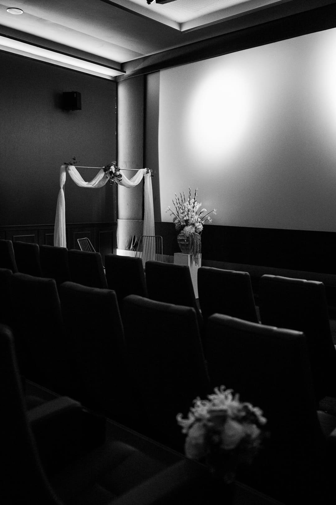 Schwarzweiß Bild eines leeren Kinosaals, vor dessen Leinwand ein Traubogen und Blumen stehen