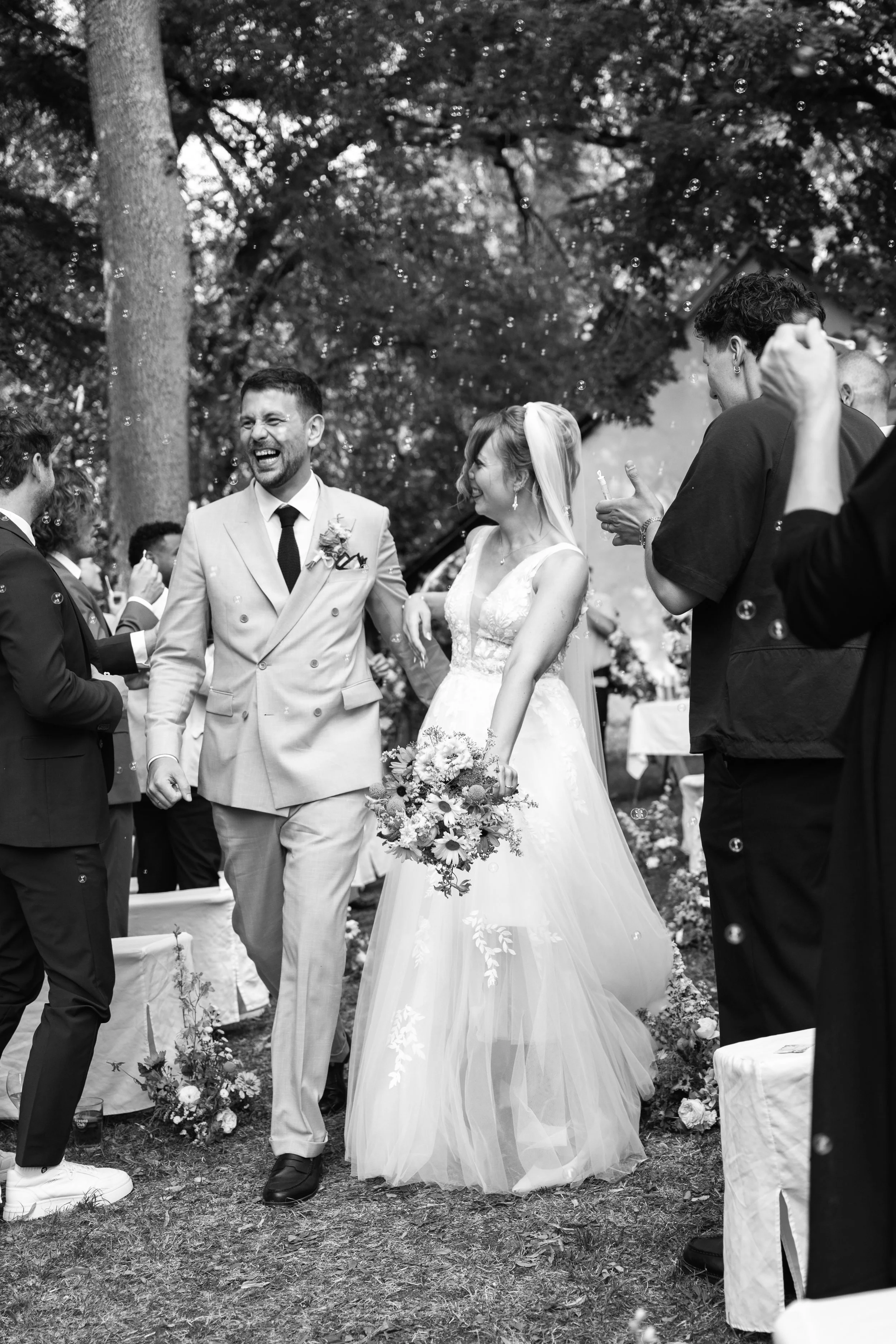 A group of people, including a smiling bride and groom, celebrating outdoors at a wedding. The bride is holding a bouquet of flowers, and guests are around them, some blowing bubbles. There are trees in the background.