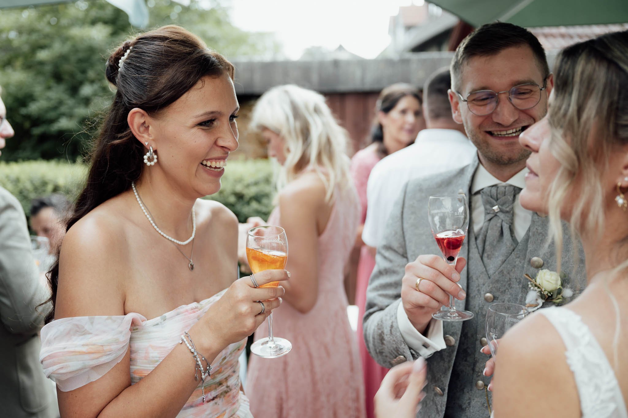 Hochzeitspaar mit Gästen beim Empfang im Garten, alle lachen und trinken Sekt.