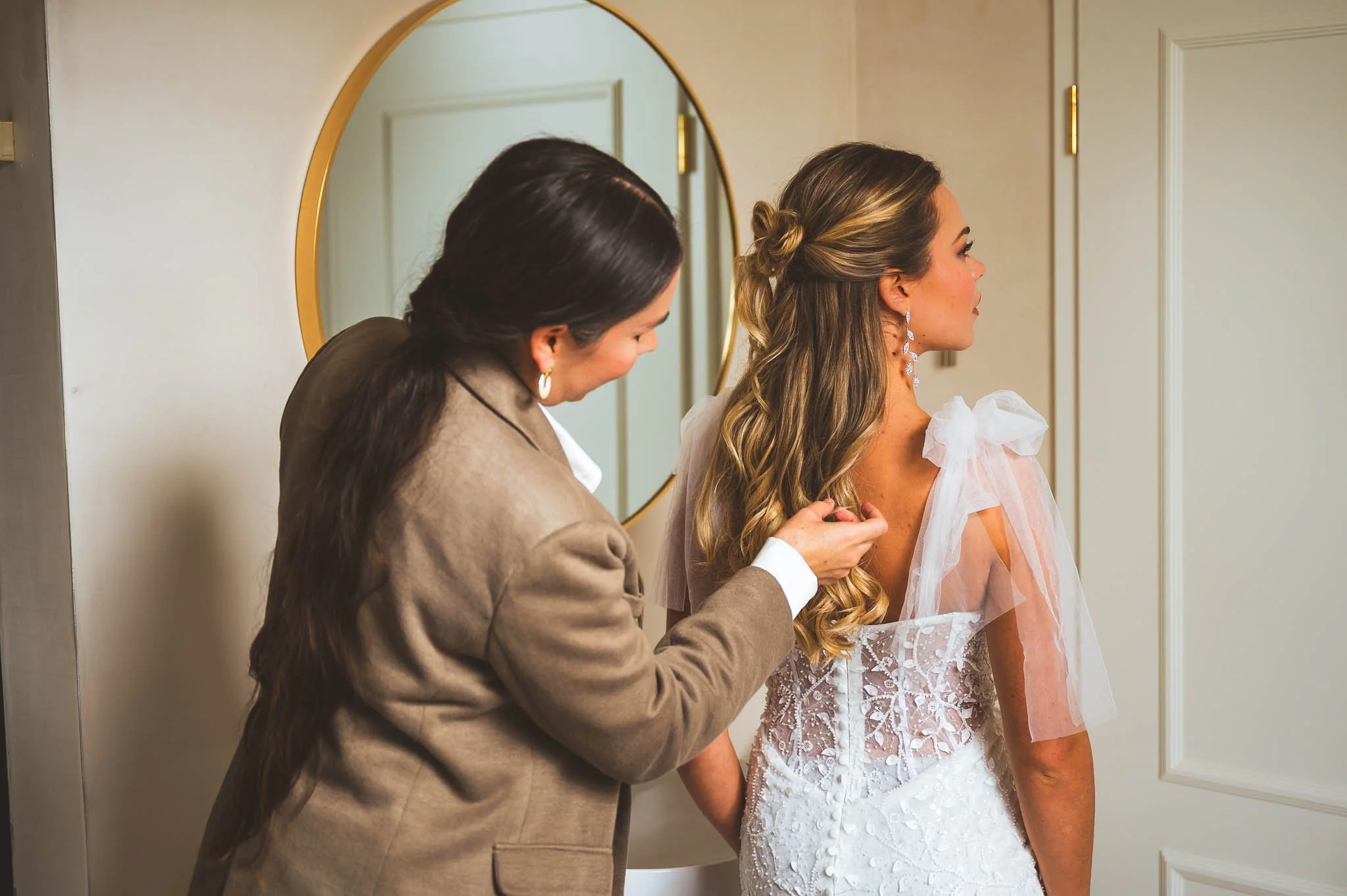 Eine Frau in einem weißen Hochzeitskleid wird von einer weiteren Frau vor einem Spiegel gestylt.