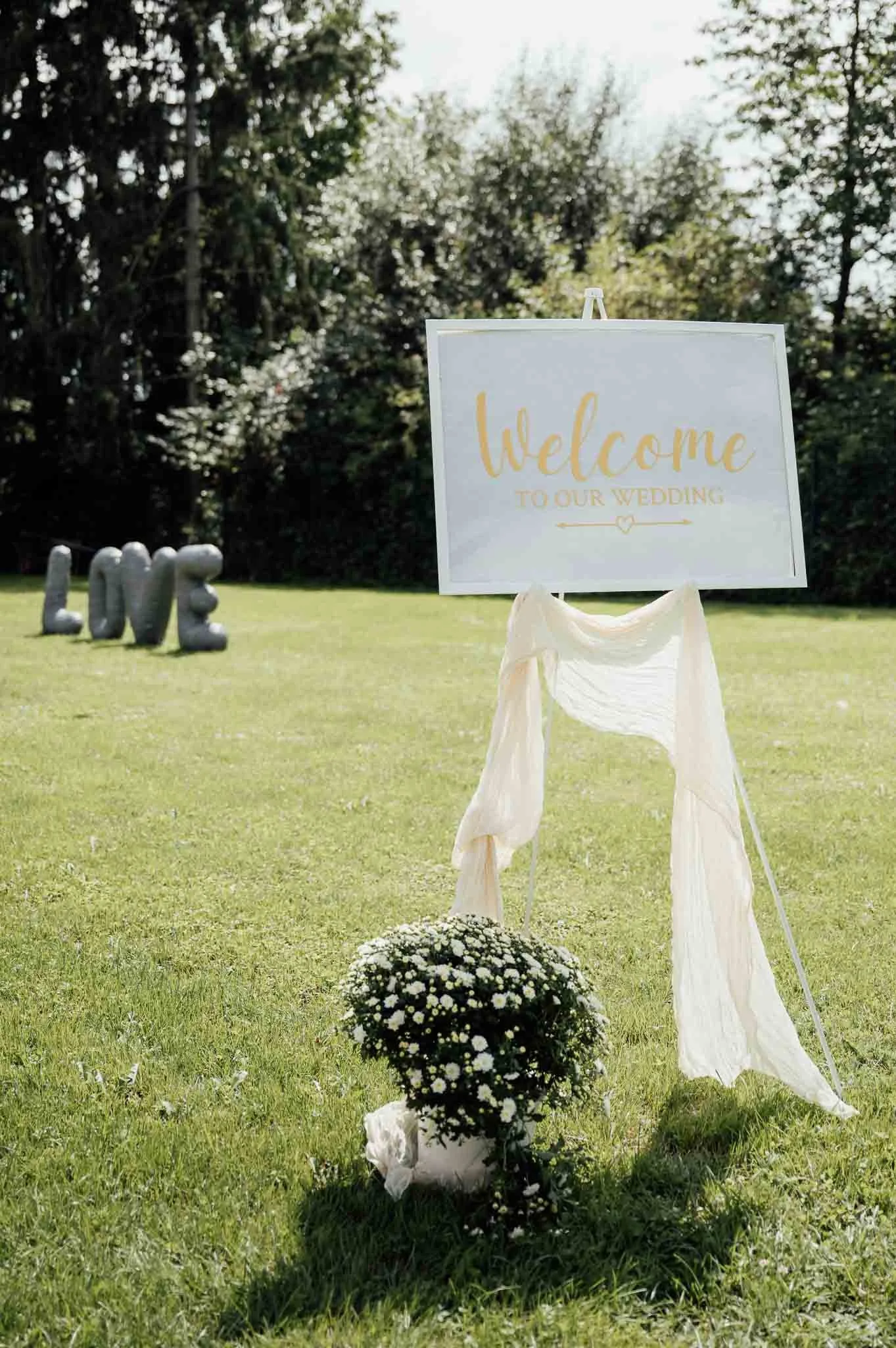 Willkommensschild für eine Hochzeit im Freien mit weißen Blumen, einem Stofftuch und löffelähnlichen Dekorationen auf dem Rasen.