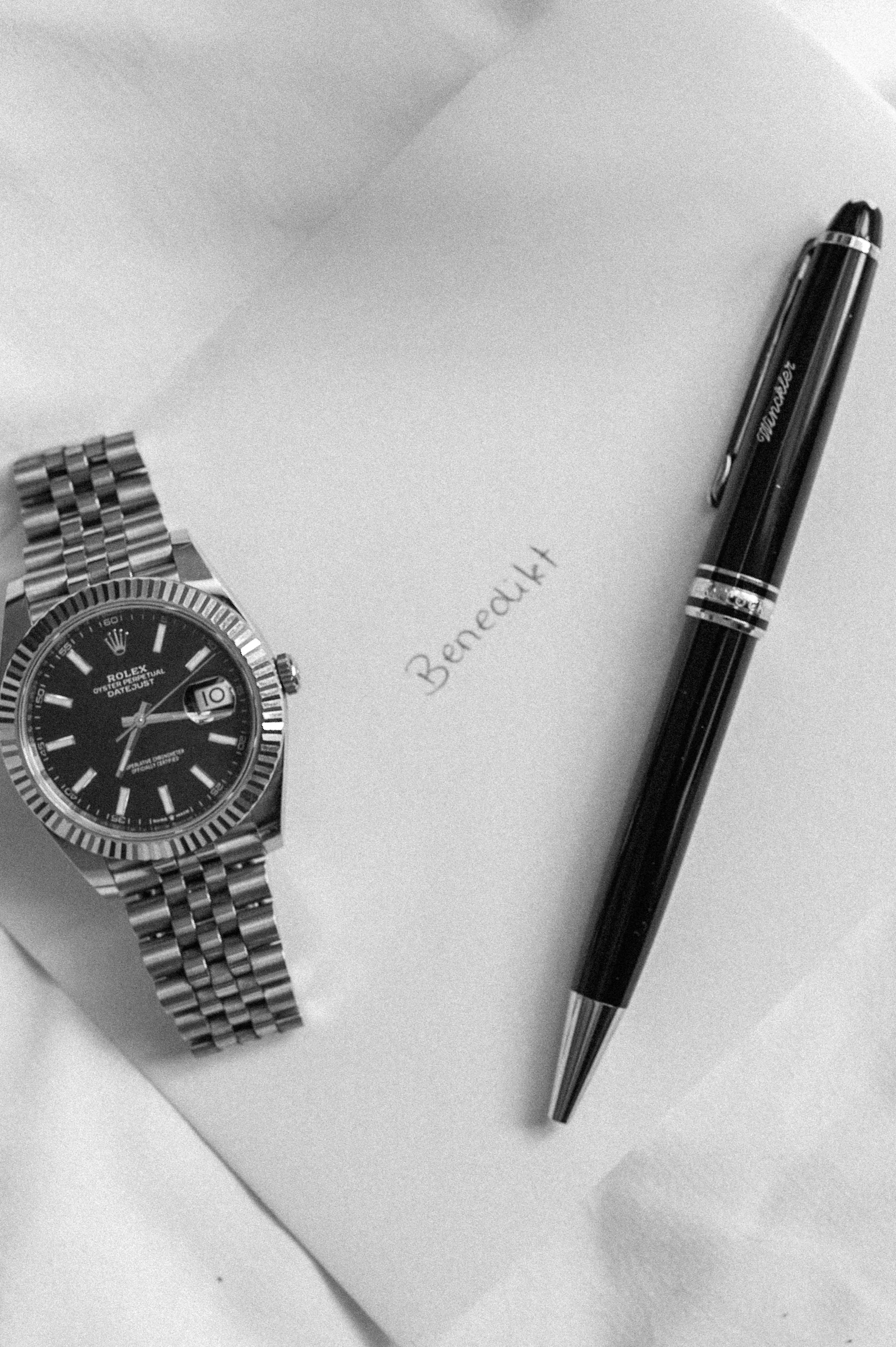 A wristwatch, a pen, a handwritten note with the name 'Bernadk', and a blank sheet of paper on a desk.