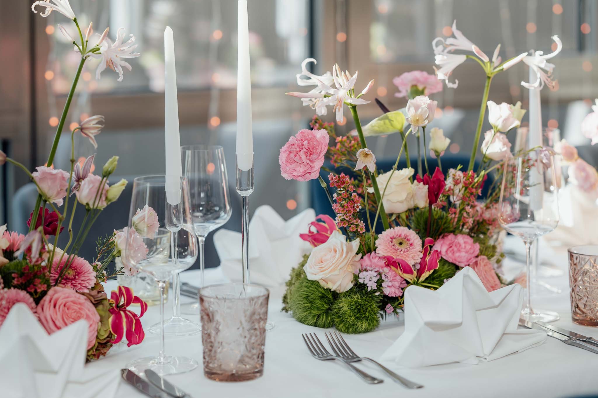Eleganter festlicher Tisch mit pinkfarbenen Blumen, weißen Kerzen, Wein- und Wassergläsern, konvexen Gläsern und weißen servietten, gedeckt für eine Veranstaltung.