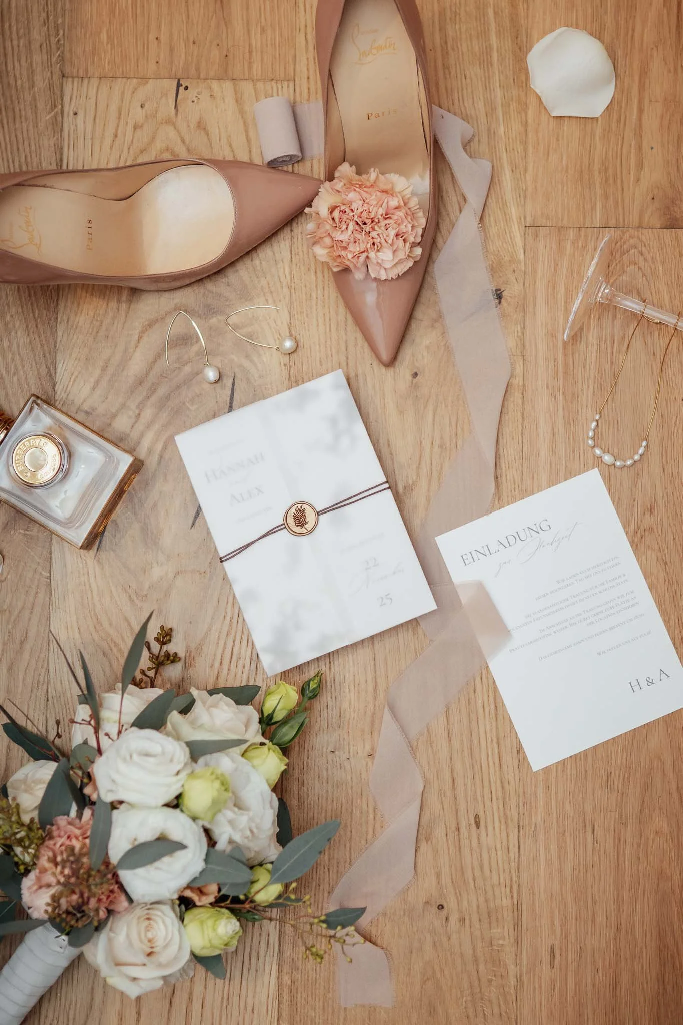 Hochzeitsdetails auf Holztisch: Brautstrauß, Einladungskarten, Pumps, Perlenohrringe, Parfümflasche, Rose, Seidenband, Lippenstift, Haarband, 2 Rosenkelche.