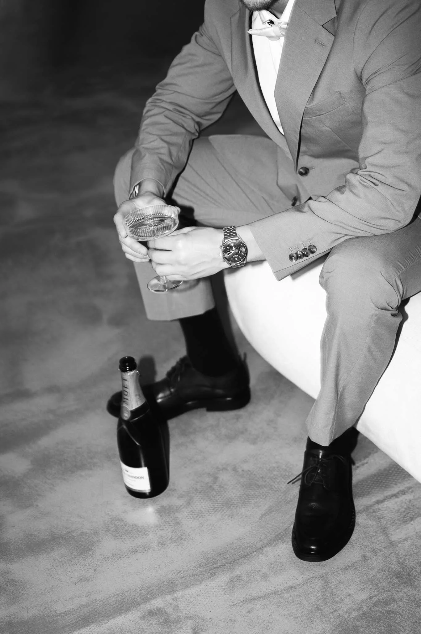 Mann in Anzug sitzt auf einem Sofa, hält ein Glas in der Hand, eine Flasche Sekt steht auf dem Boden.