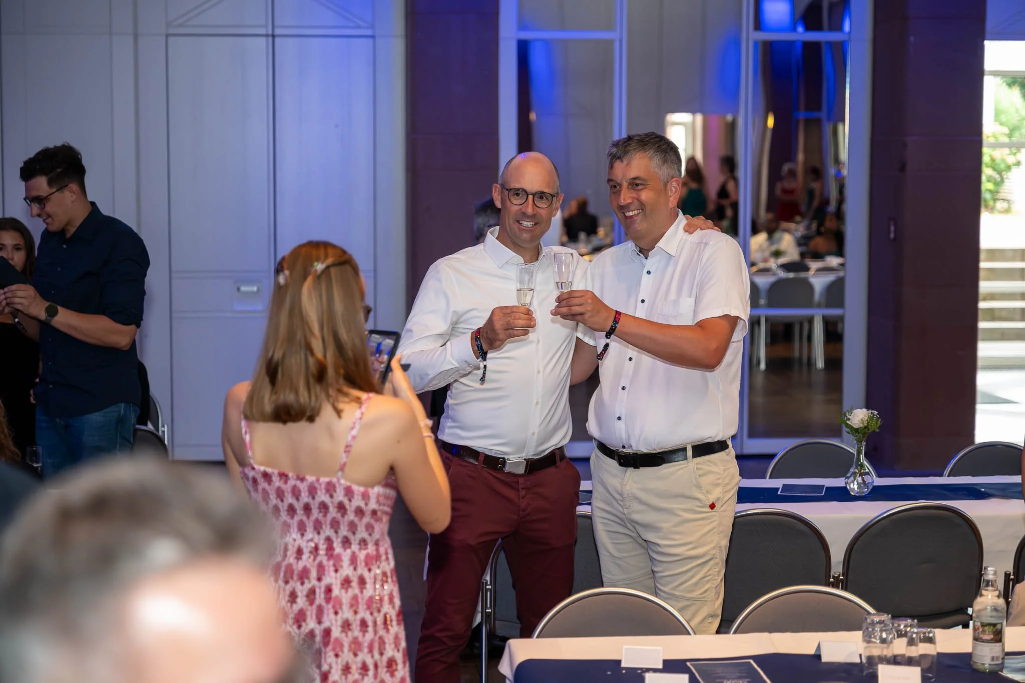 Zwei Männer stoßen auf einer Dinnerparty an, umgeben von festlicher Atmosphäre und fröhlichen Gästen
