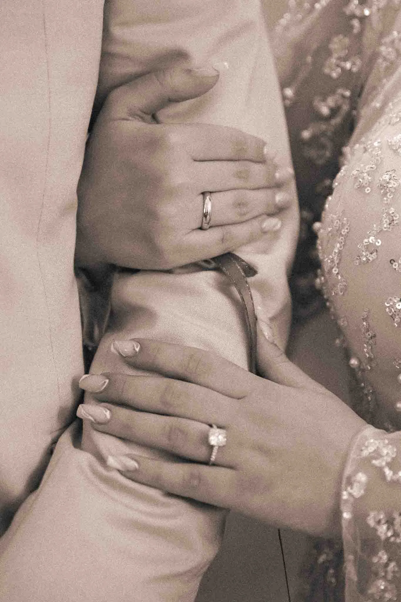 Hände eines Paares mit Eheringen, die auf einem Arm ruhen, eine trägt einen großen solitaire Diamantring, die andere einen einfachen Ehering. Die Frauen trägt ein Kleid mit Pailletten und Seide, das Männer trägt einen Anzug.