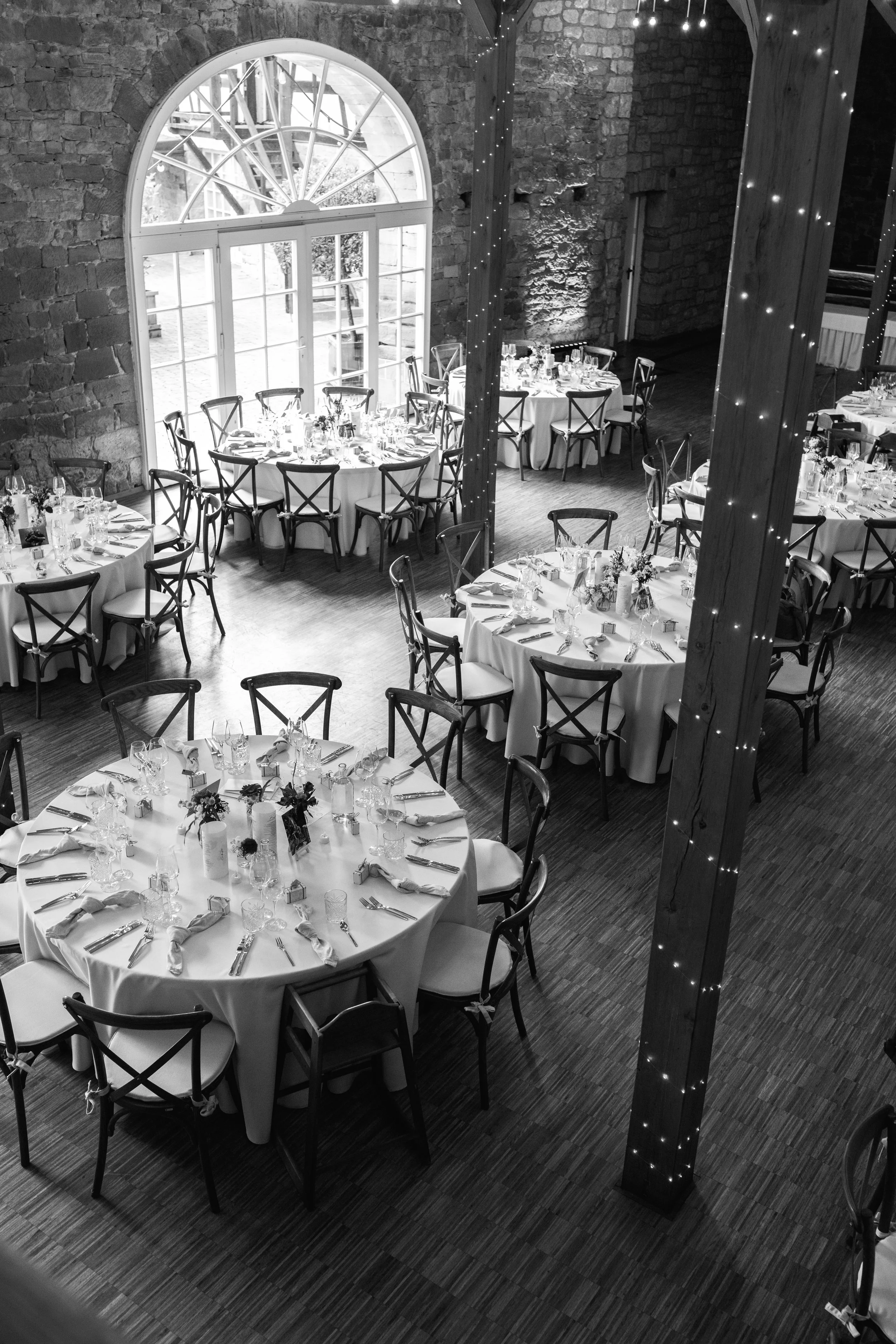 Indoor wedding reception with round tables covered in white tablecloths, set with glasses, napkins, and floral centerpieces. The venue has large arched windows, exposed brick walls, and wooden beams decorated with string lights.