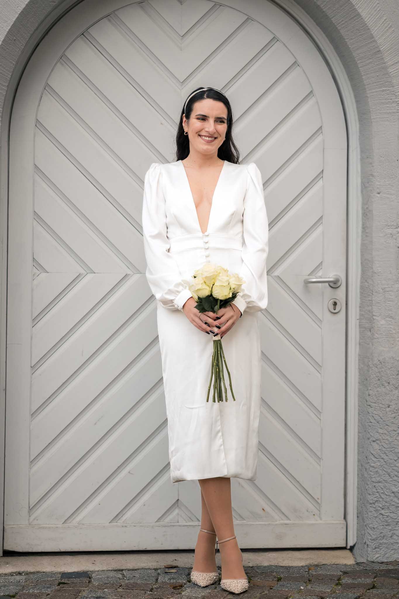 Eine Braut steht in einem weißen Kleid und einem Blumenstrauß vor ihm Bauch vor einer alten Türe und lächelt, während sie in die Ferne blickt