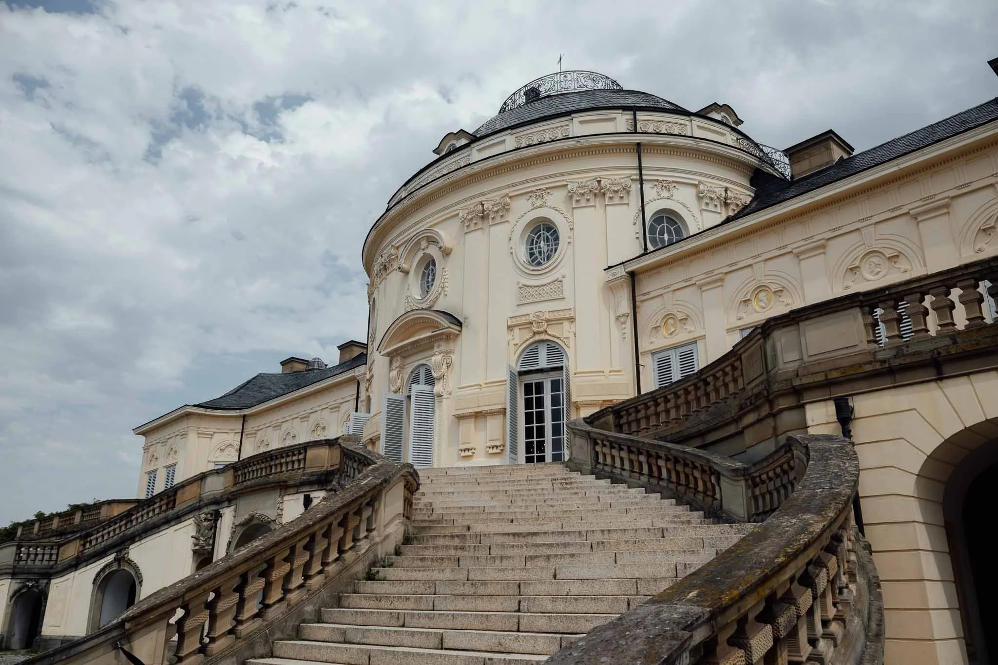 Ein barockes Gebäude mit einer Treppe aus Stein, das auf einen Hügel führt. Die Fassade ist cremefarben mit dekorativen Elementen und mehreren runden Fenstern. Der Himmel ist teilweise bewölkt.