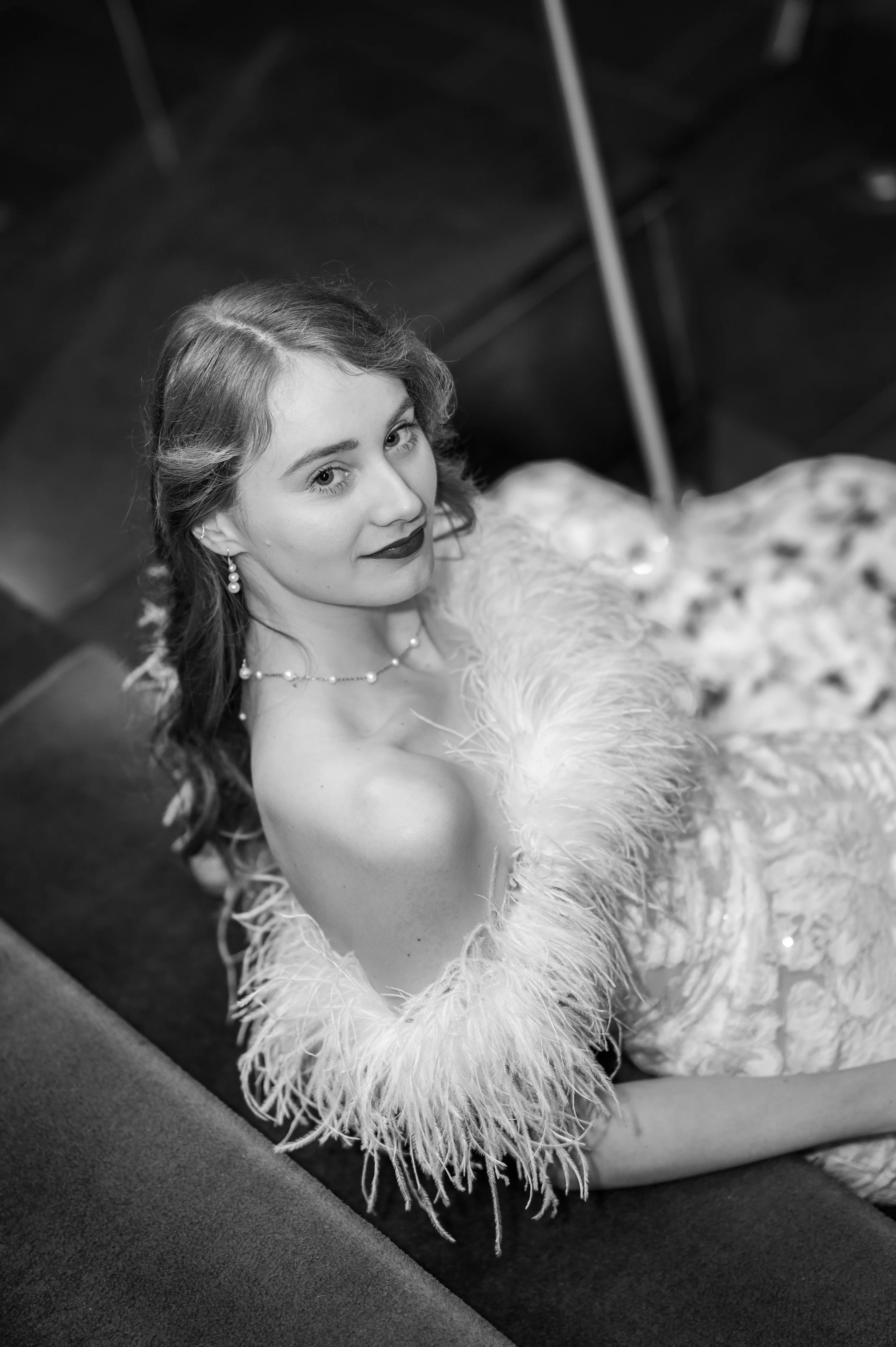 Schwarzweiß Bild einer Frau in einem extravaganten Kleid mit Federn am Ausschnitt. Sie sieht direkt in die Kamera und das Bild wirkt sehr dramatisch und glamourös. Sie trägt die welligen Haare offen, ist dezent geschminkt mit dunklen Lippen