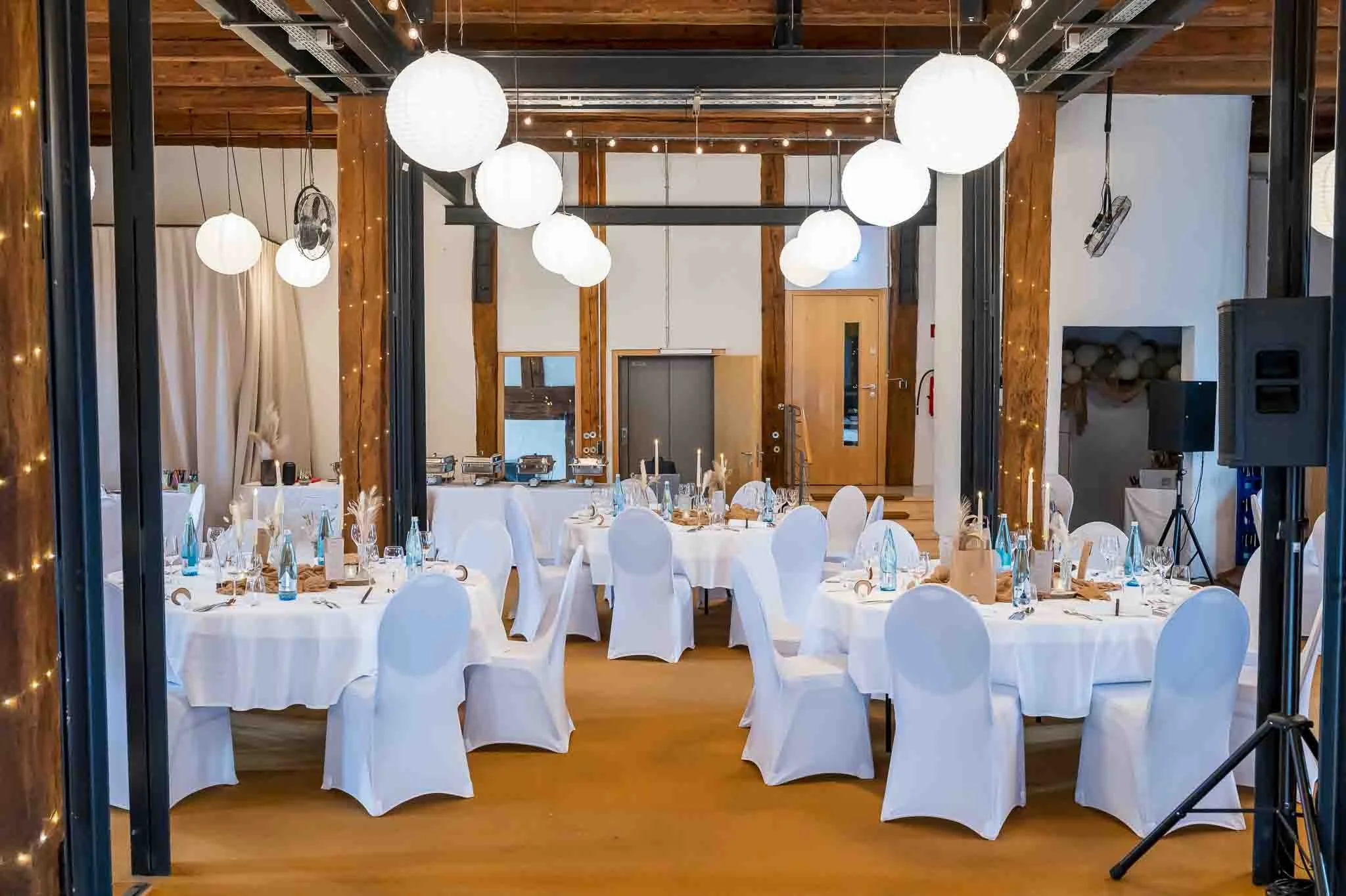 Bild eines gedeckten Festsaales in einer scheunenartigen Location. Zu sehen sind Stühle mit weßen Stuhlhussen und runde Tische mit blauen Glasflaschen darauf