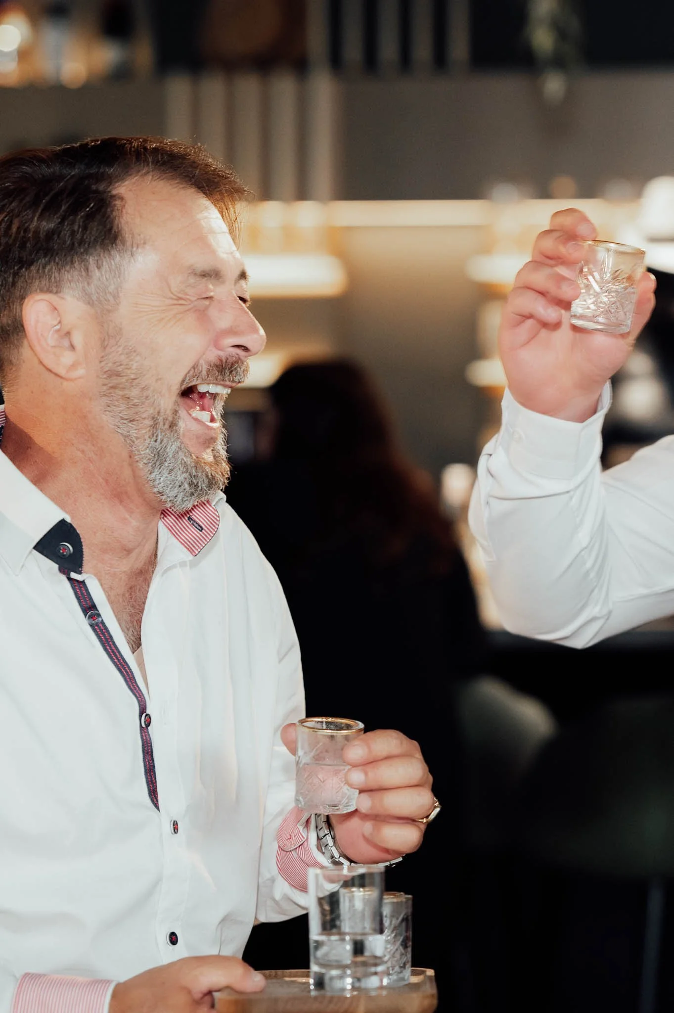 Ein lachender Mann mit grauem Bart hebt ein Glas mit Whisky in einem Café oder Restaurant.