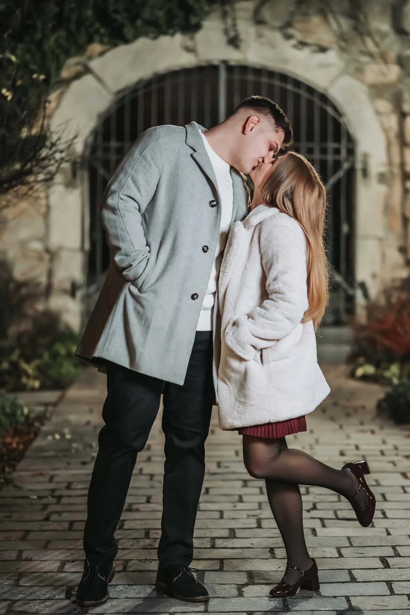 Ganzkörperaufnahme von einem Paar, welches sich vor einem Tor an einer Steinmauer küsst. Sie hebt dabei ein Bein hoch. Beide tragen Mäntel und es ist nacht.