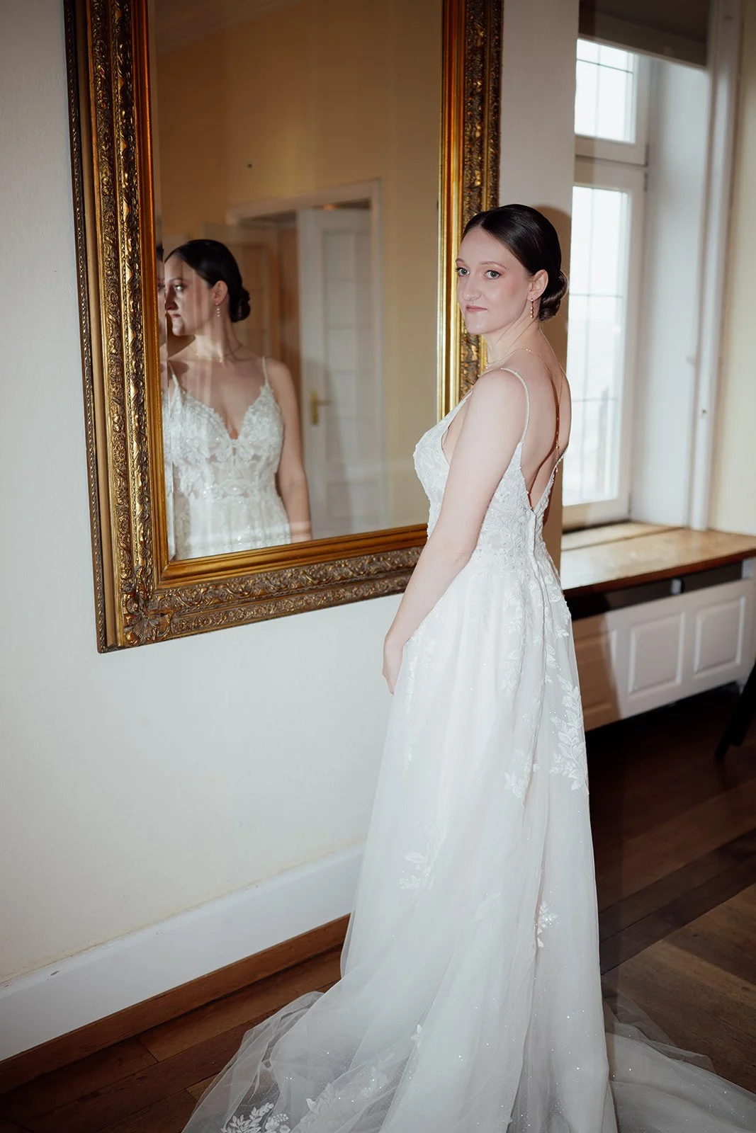 Bild einer Braut, die vor einem barocken Spiegel steht. Sie blickt halb ernst über ihre Schulter an der Kamera vorbei