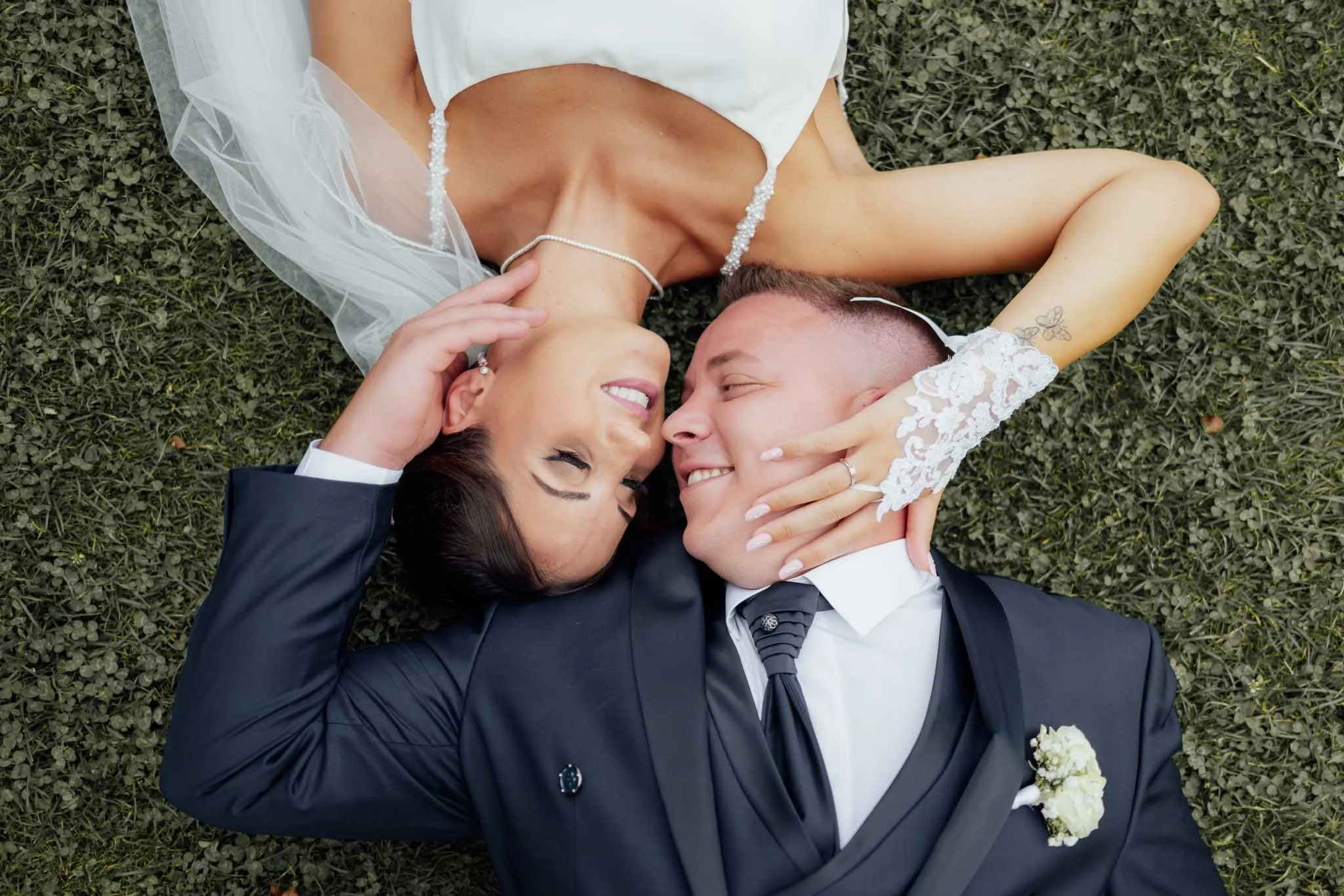 Ein verliebtes Paar liegt auf dem Rasen, die Frau trägt ein weißes Brautkleid und die Haare sind offen. Der Mann trägt einen schwarzen Anzug mit Krawatte. Sie lachen und schauen sich liebevoll an.