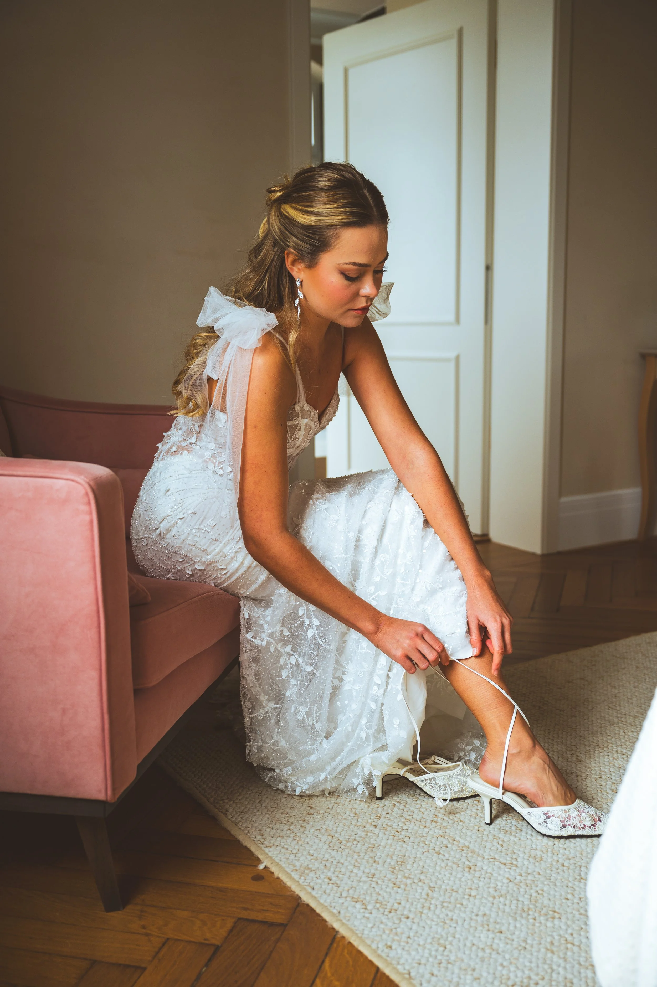 Eine Braut sitzt auf einem rosa Sessel und schließt ihren Schuh